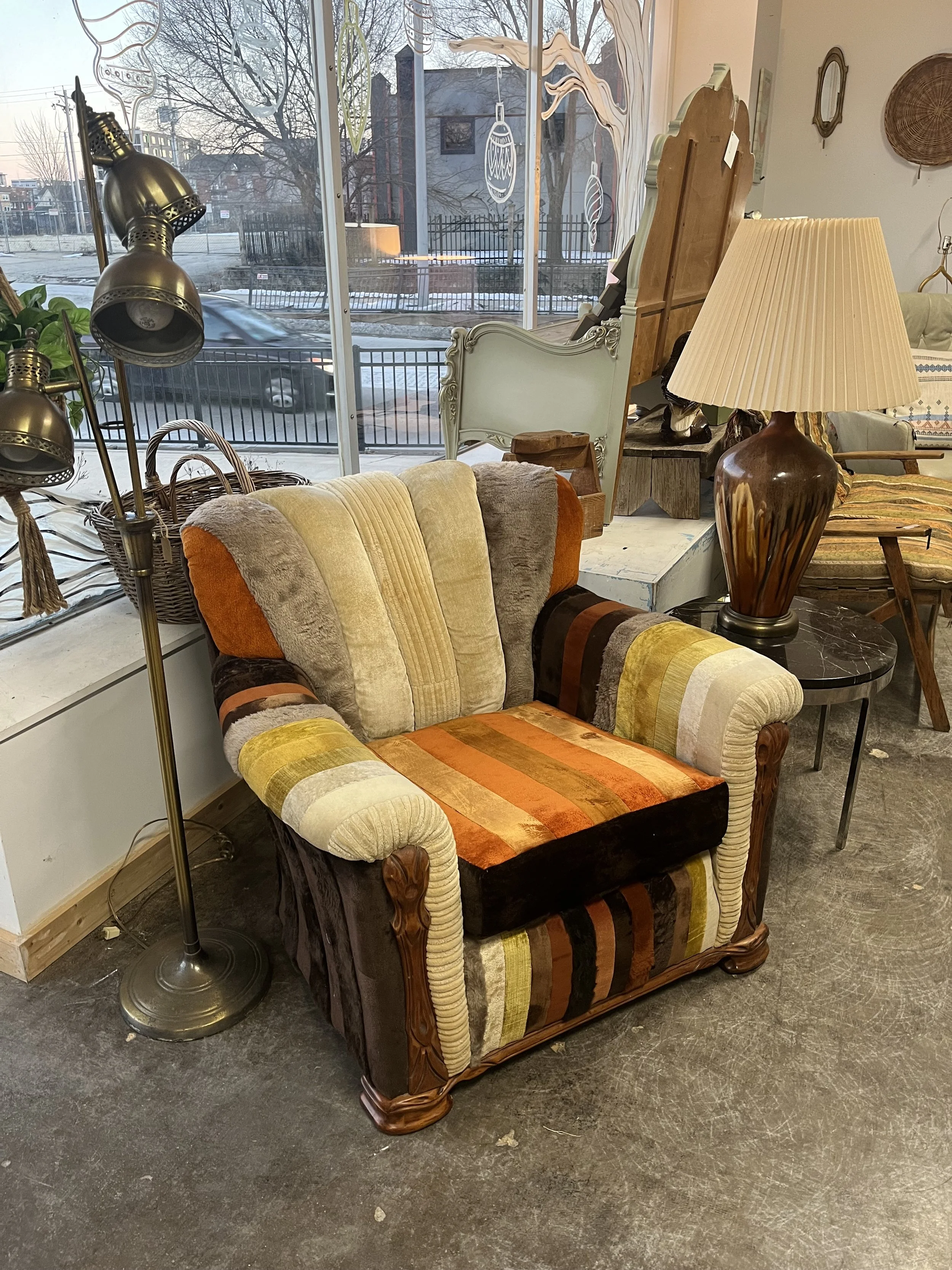 Colorful vintage armchair with striped fabric in shades of orange, yellow, brown, and beige next to a wooden side table with a large ceramic lamp, inside a furniture store window, with a snowy outdoor scene visible through the window.