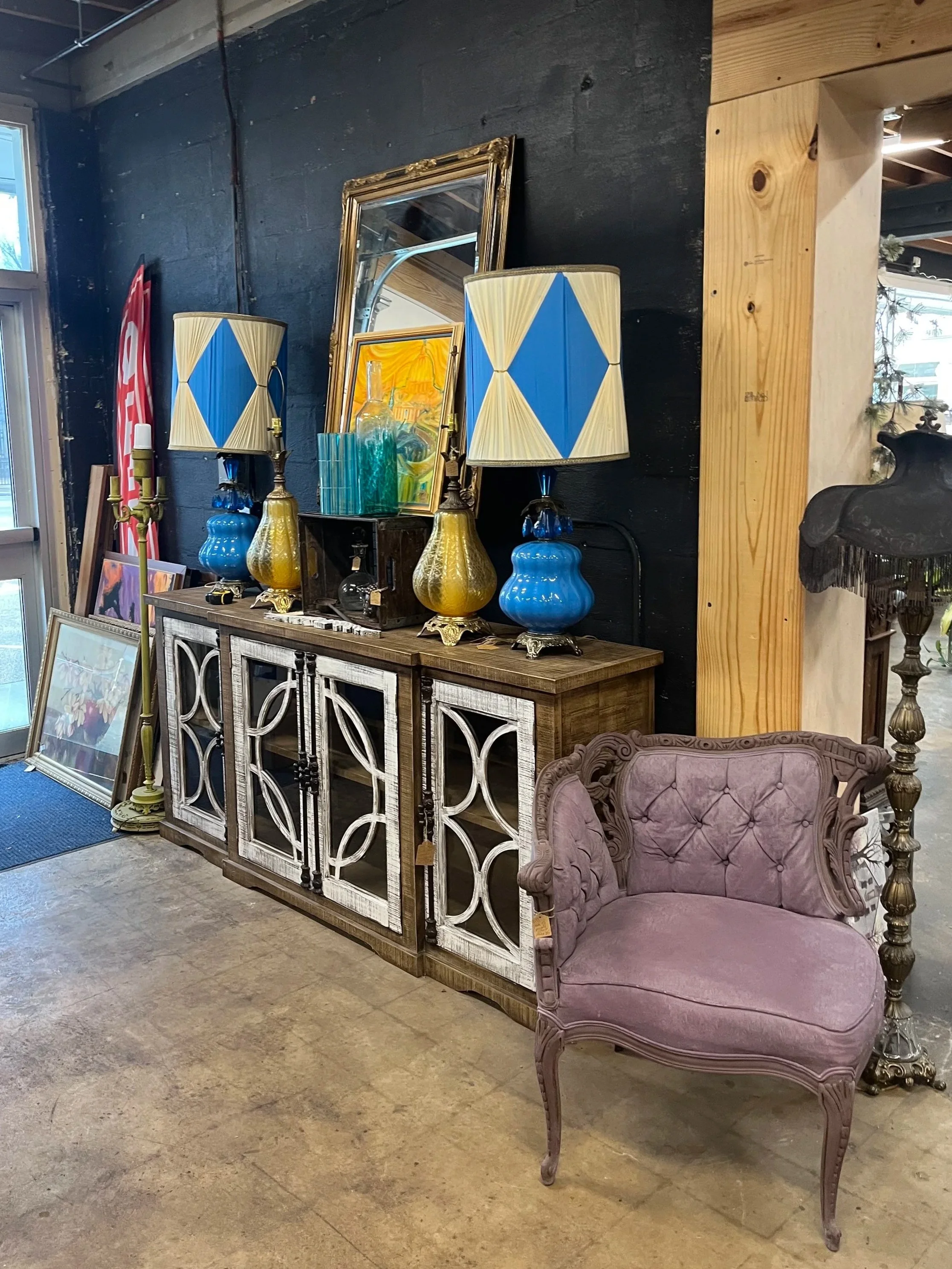 A vintage-style purple velvet armchair with carved wooden details next to a wooden lamp with a black shade and brass stand, against a dark wall with a wooden beam.