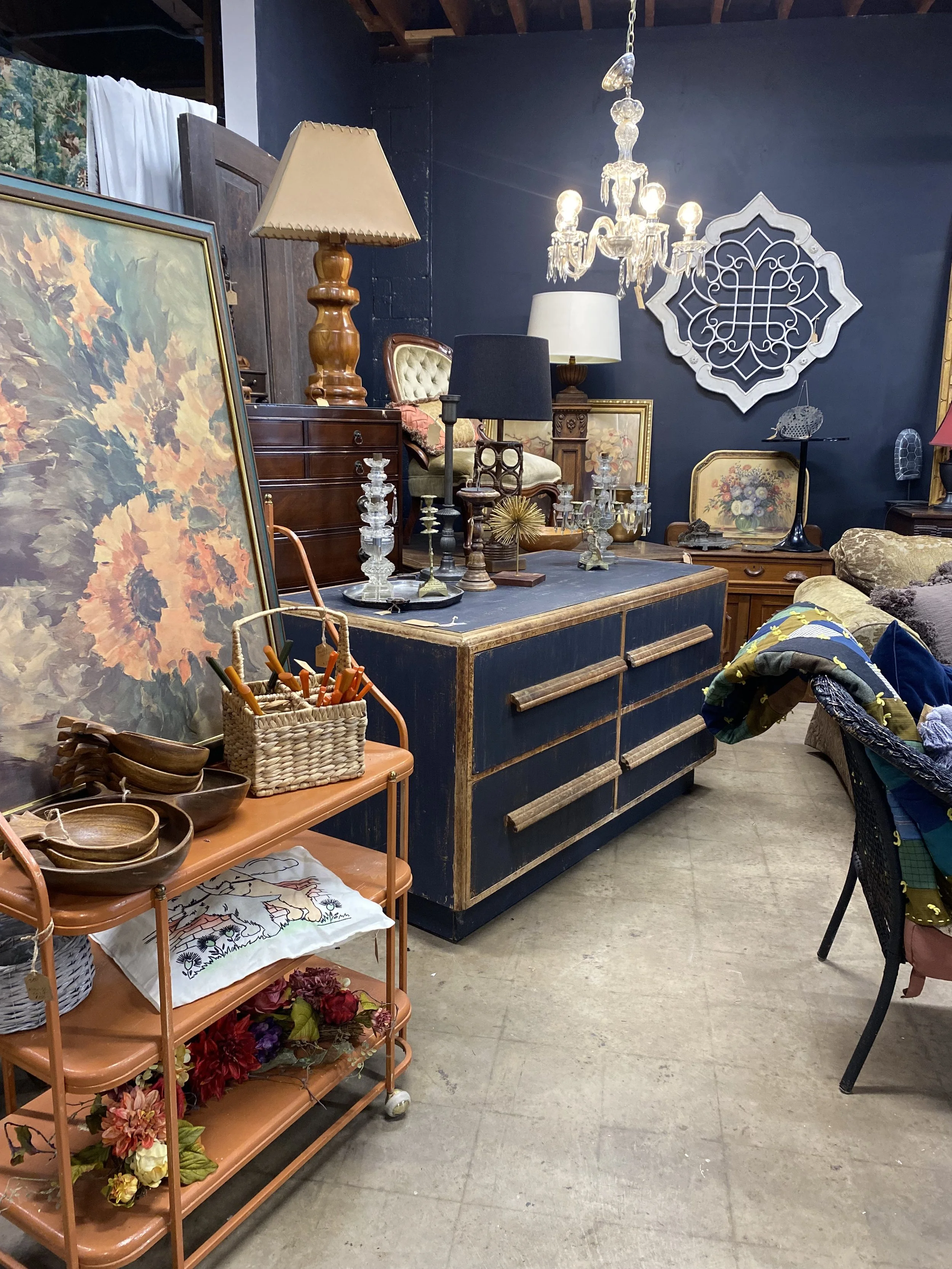 An antique and vintage furniture store with a variety of decorative items, including a large sunflower painting, glass candlesticks, table lamps, a chandelier, a wooden dresser, and a basket with utensils on a cart.