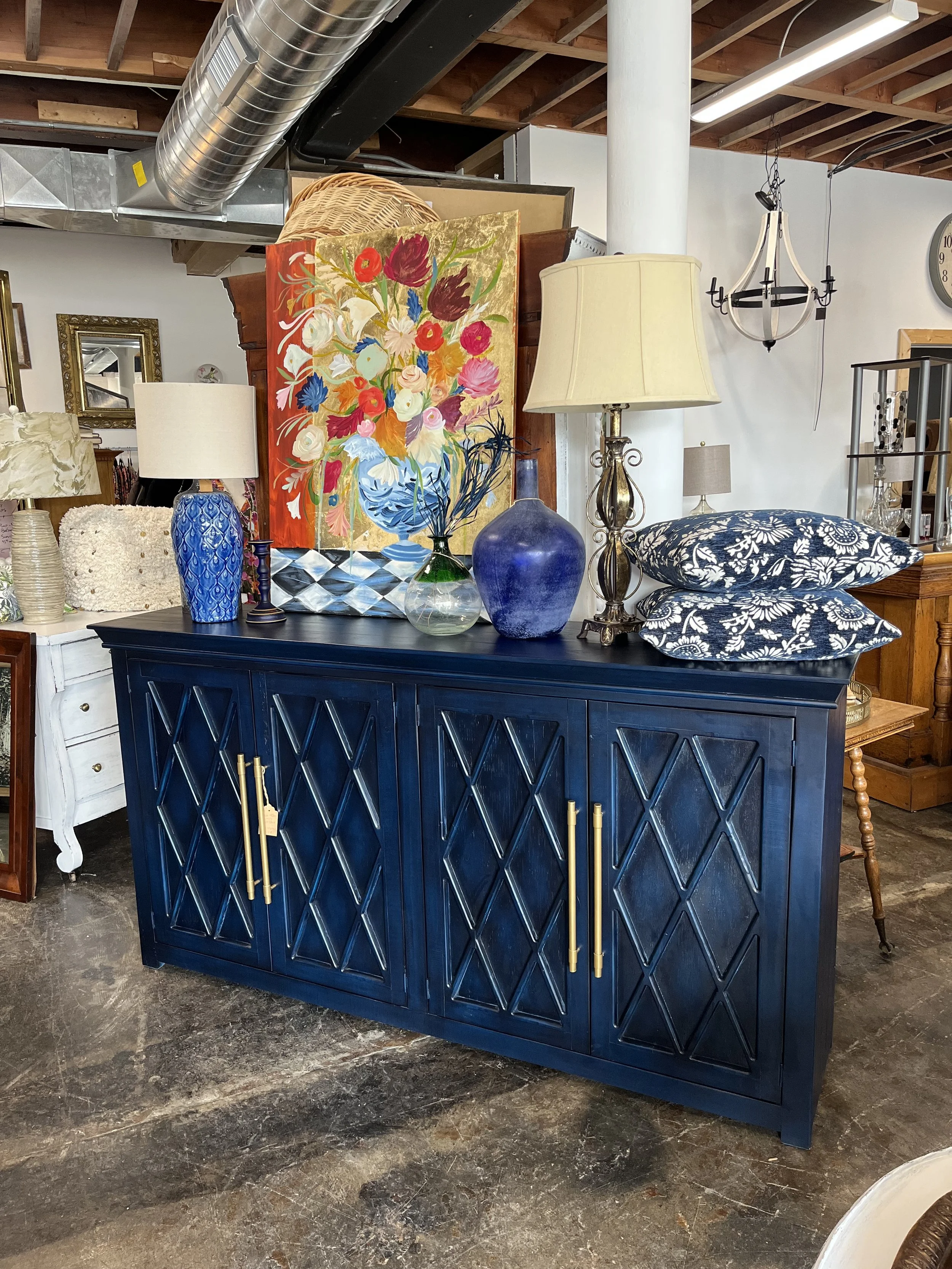 A dark blue sideboard with gold handles, decorated with colorful vases, a table lamp, and two navy and white patterned pillows, set in an antique or thrift store.