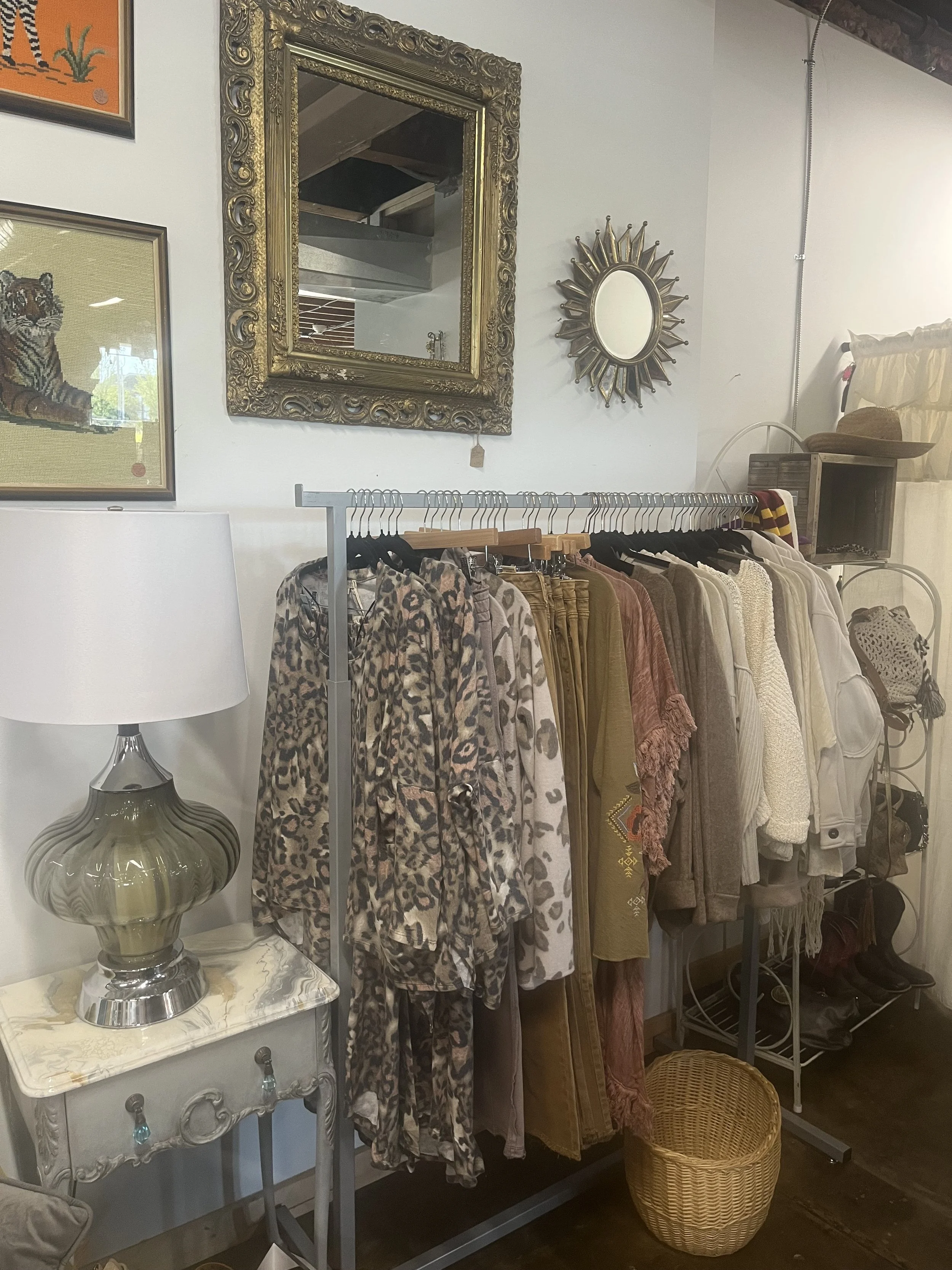 Clothing rack with animal print, beige, brown, and cream-colored clothes, a decorative lamp on a side table, and various wall mirrors and art on the wall.
