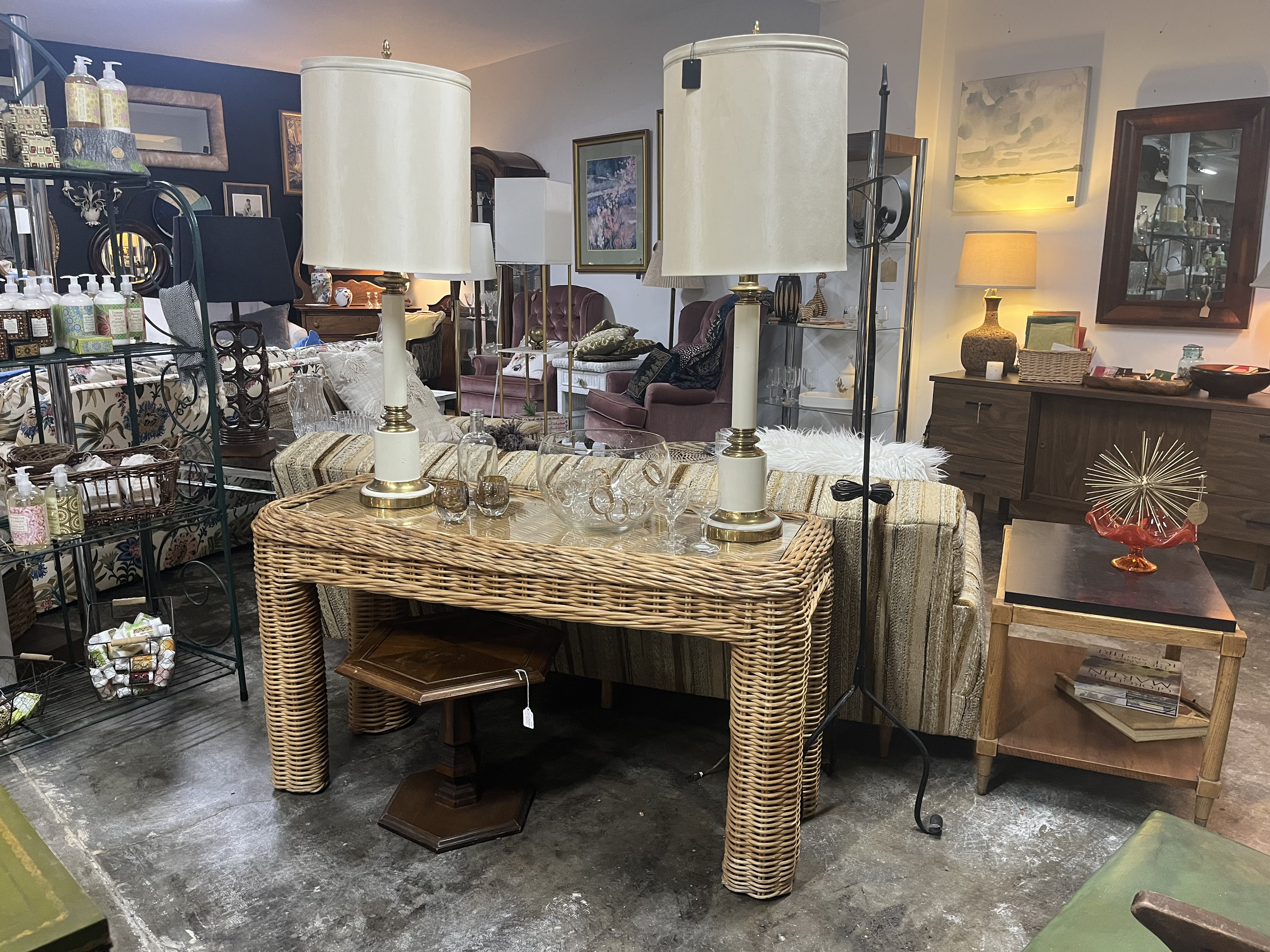 Furniture and decor in an antique or thrift store, including lamps, tables, framed artwork, shelves with bottles and decorative items, and various other home accessories.