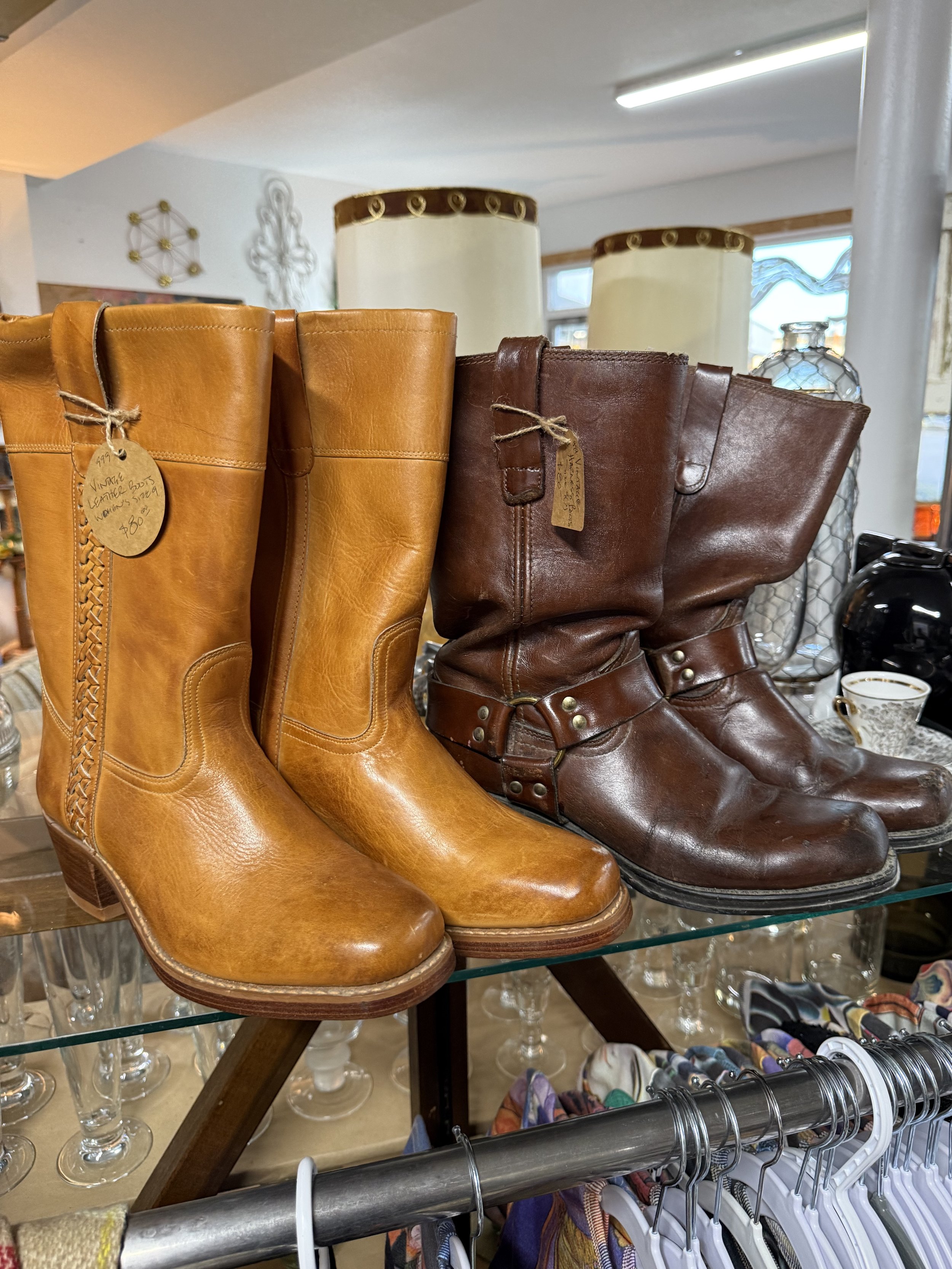 Vintage leather boots, light brown on the left and dark brown on the right, displayed on a glass shelf in a thrift store.