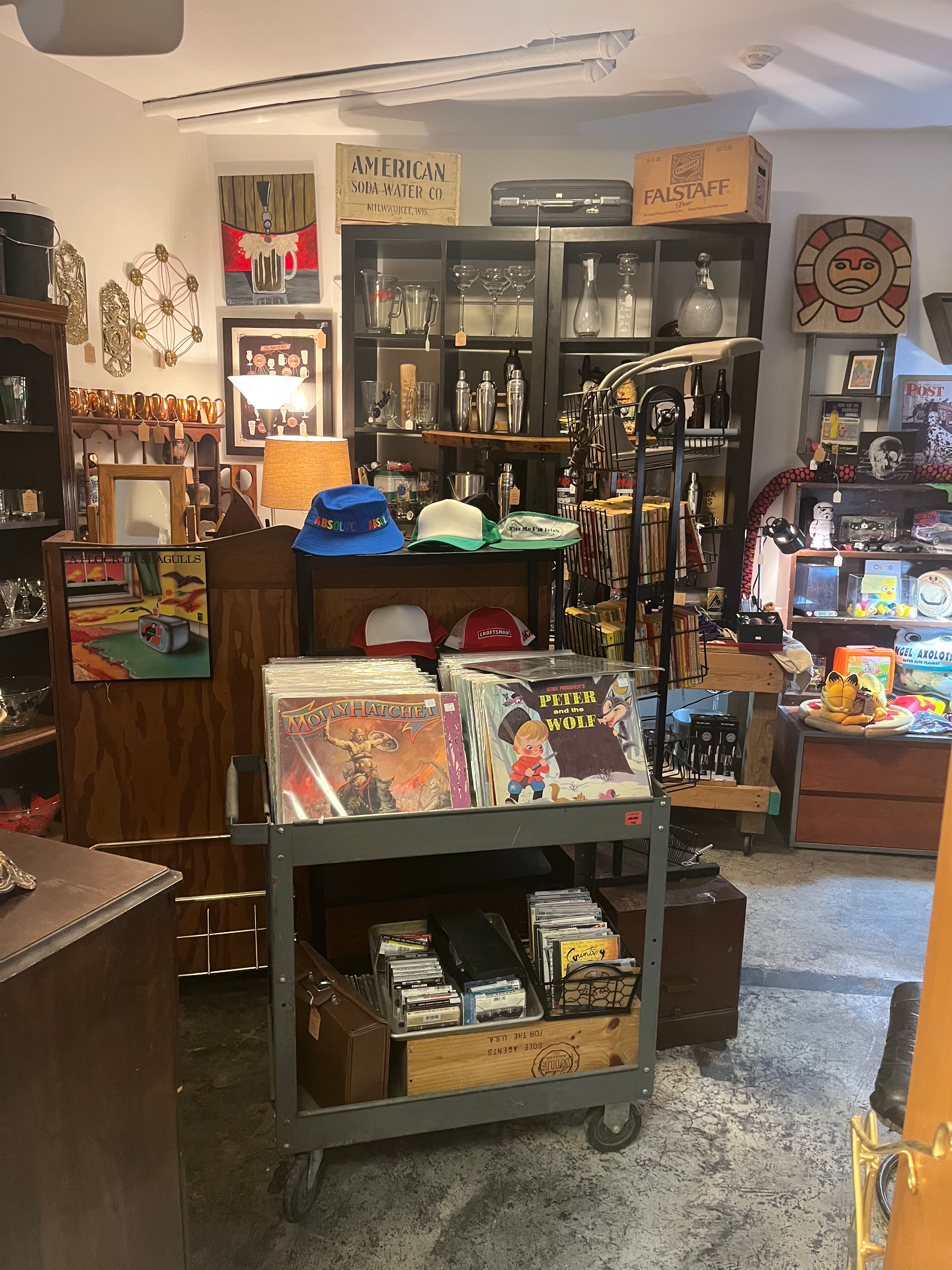 Imaginary room with shelves, books, and collectibles. A cart in the center displays comic books and cassette tapes. Hats and lamps are on the shelves and a wooden crate on the floor contains more items. Decorative art pieces and vintage items are on 