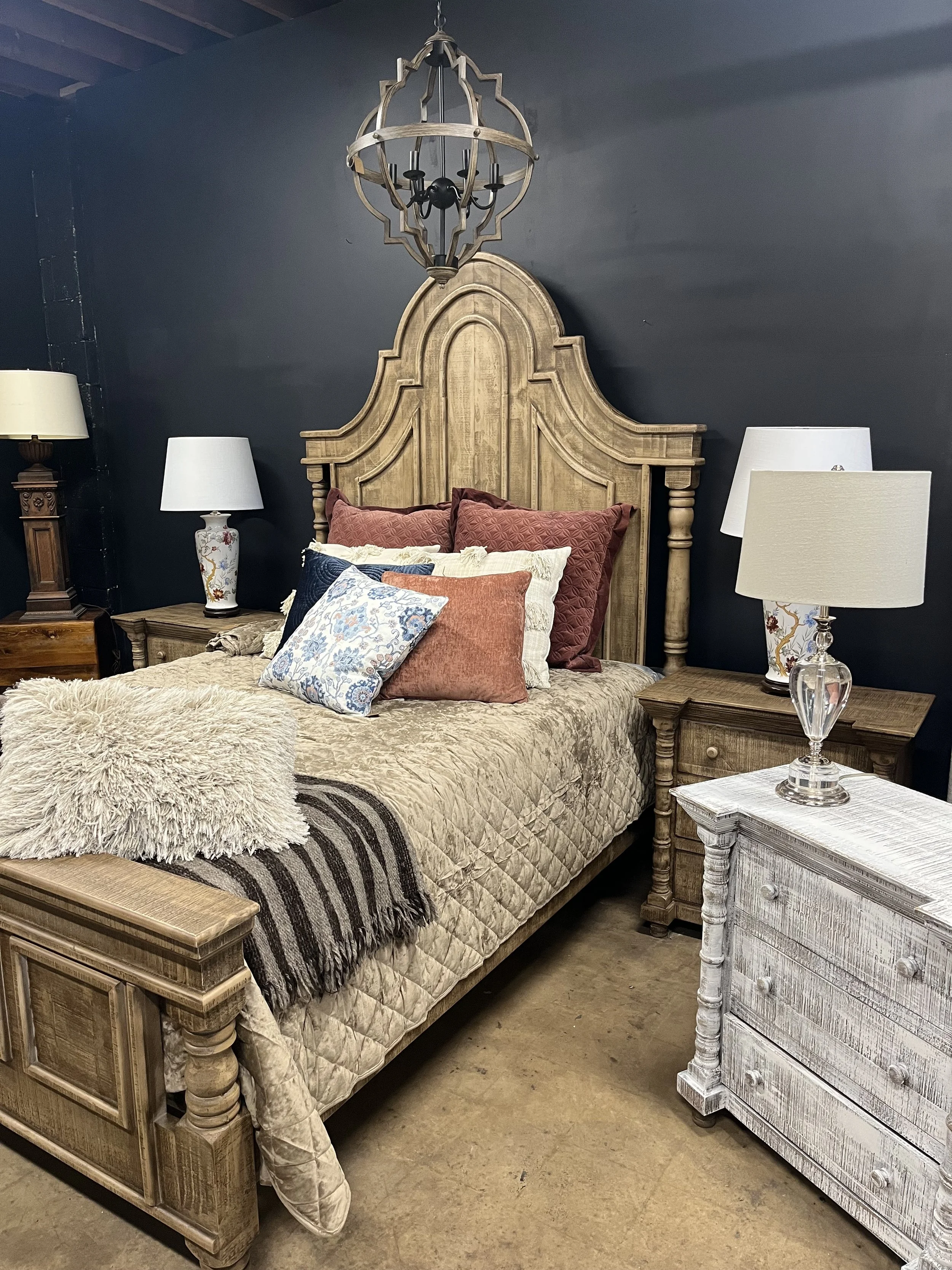 A bedroom with a large wooden headboard, patterned bedspread, and multiple pillows. There are two nightstands with lamps and a white distressed chest of drawers. A rustic chandelier hangs above the bed, and the wall behind is painted dark gray.
