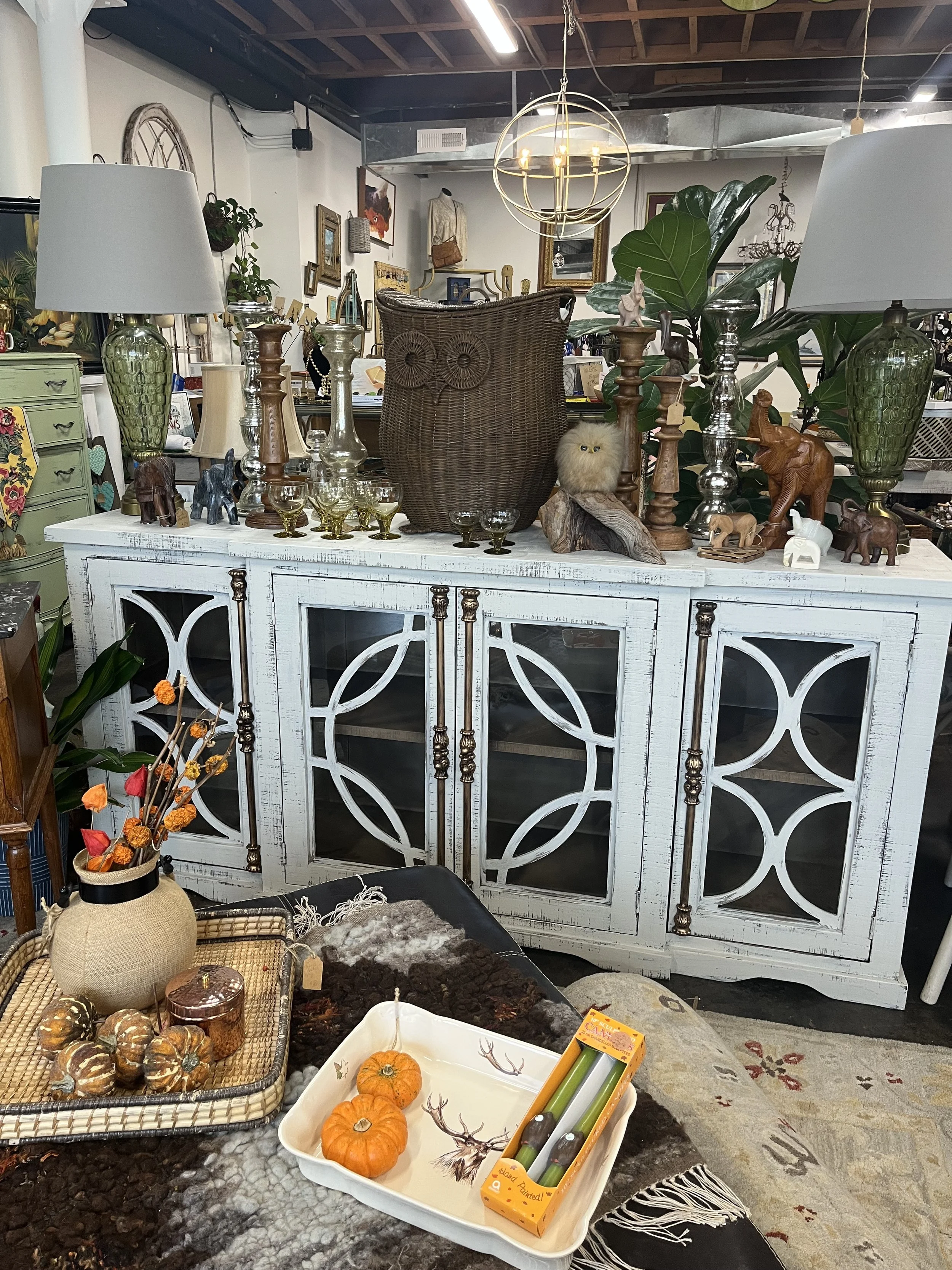 A vintage white cabinet with glass panels and decorative metal accents holds various decorative items including green and brown lamps, small elephants and other figurines, and large candles. In front, a table displays small pumpkins, dried flower arr