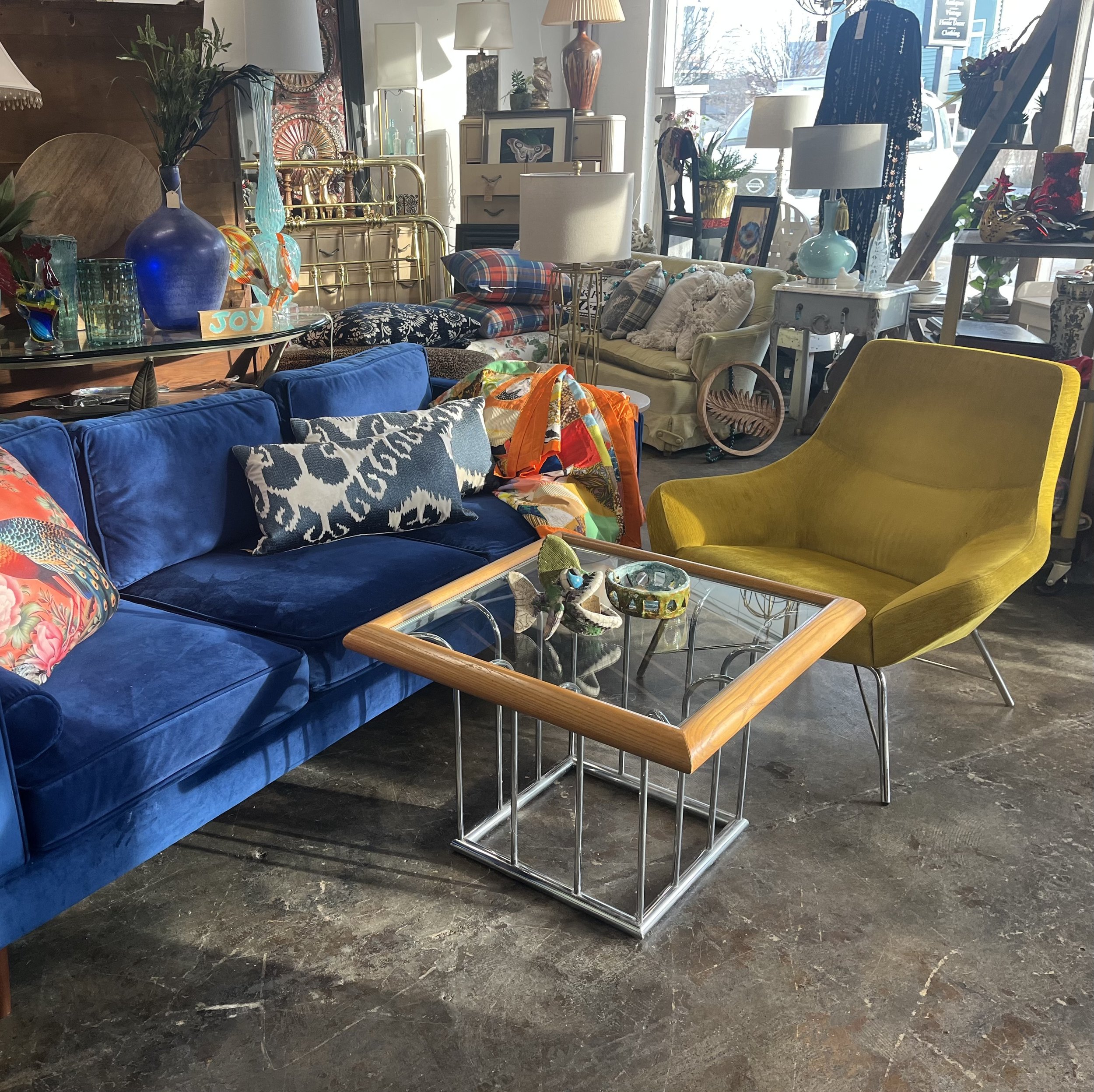 An interior view of a vintage furniture store. The foreground features a blue velvet sofa with patterned cushions, a glass-topped coffee table with decorative items, and a mustard yellow mid-century modern chair. The background displays various lamps