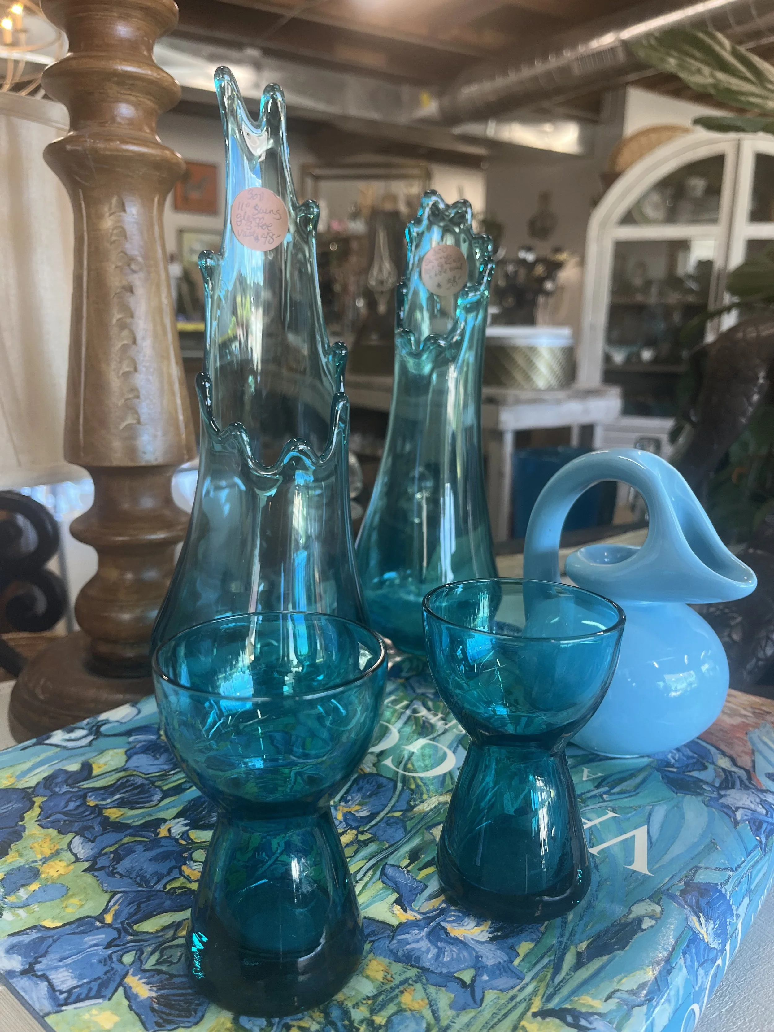 Set of three tall, blue glass vases with wavy, irregular edges, two matching teal glass goblets, and a white ceramic swan-shaped vase, all arranged on a colorful flower-patterned tablecloth in an antique shop.