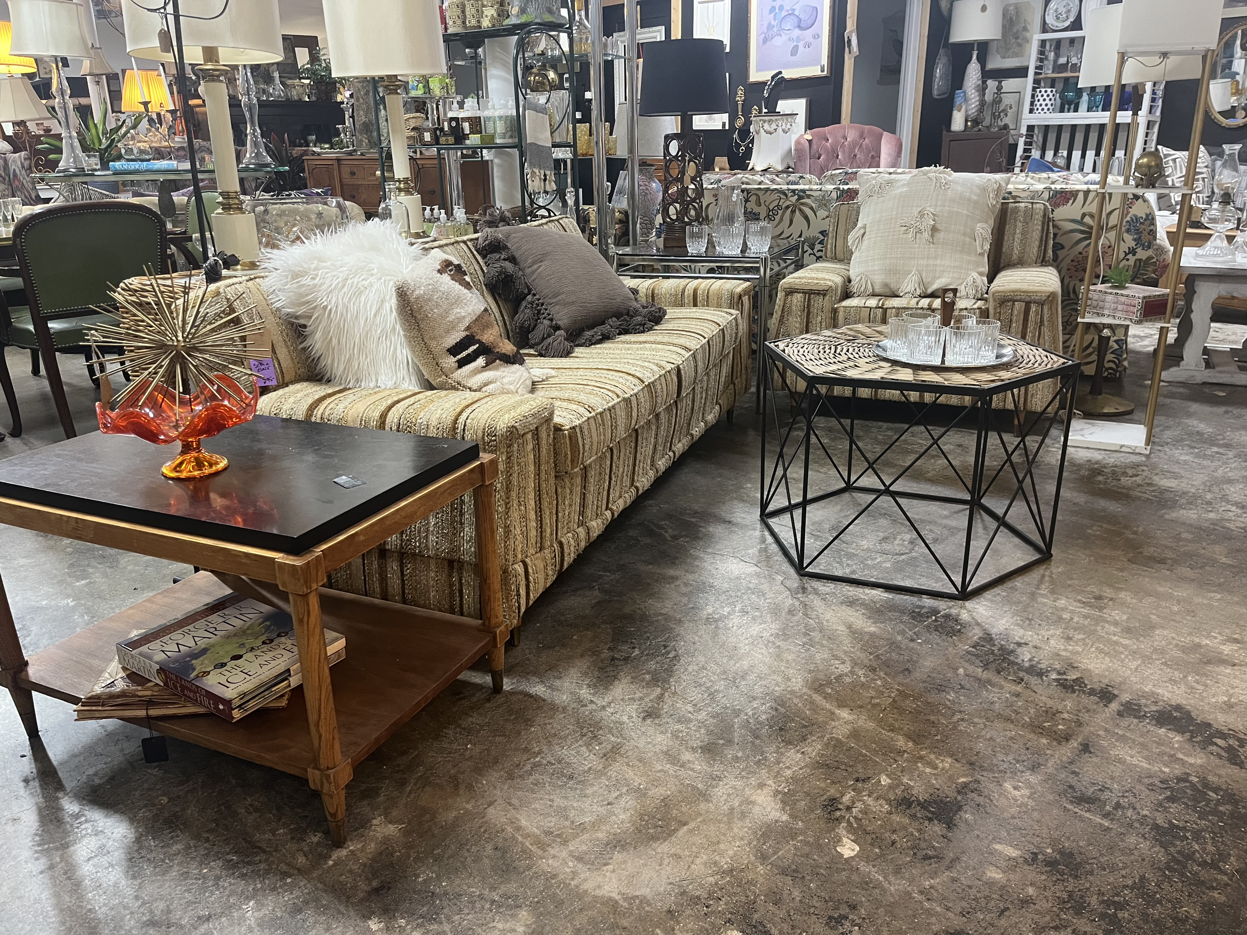 Living room setup with striped sofa, accent pillows, black coffee table, and side table with books, surrounded by other furniture and decor items in a furniture store.