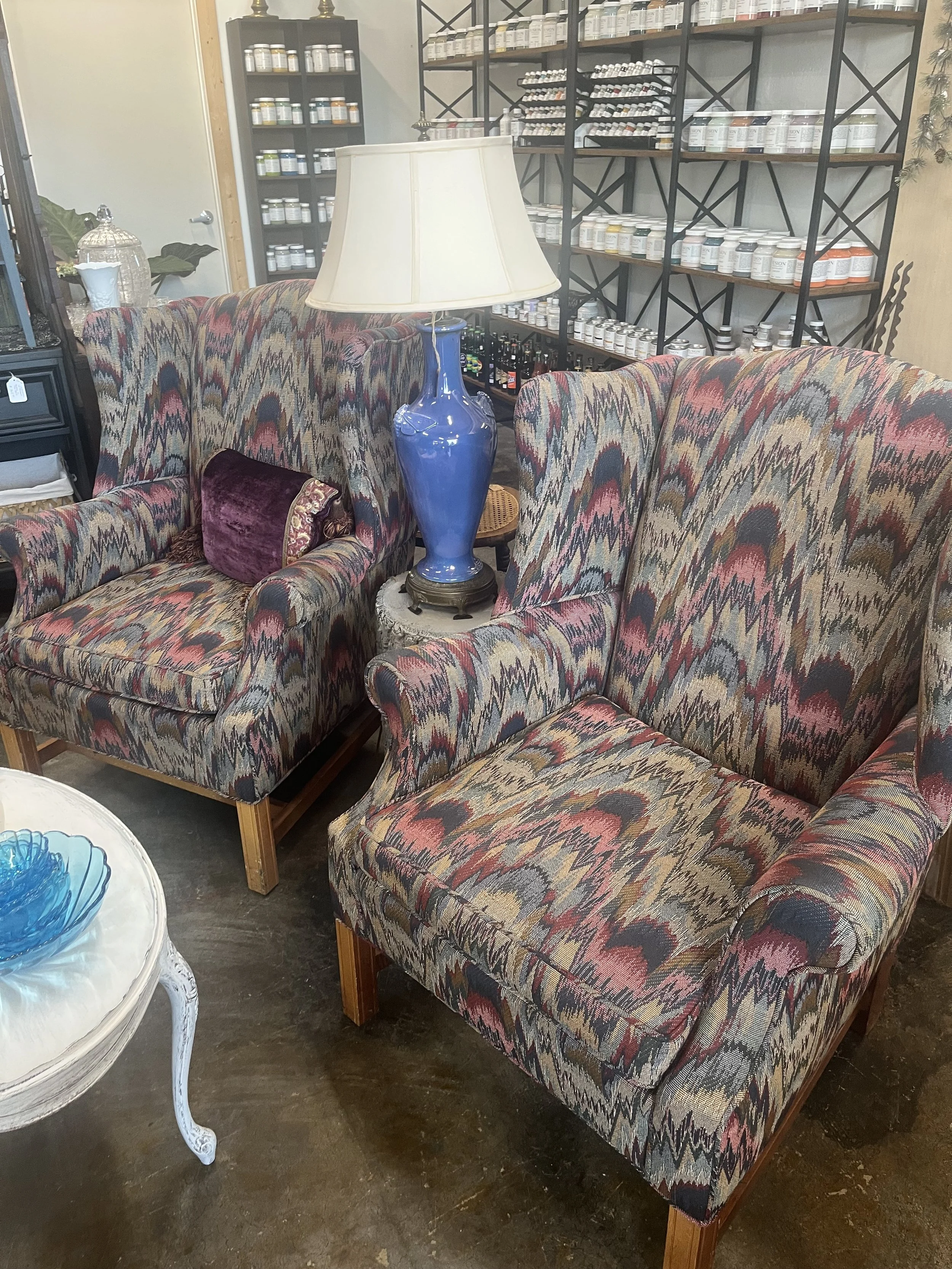 Vintage armchair with colorful zigzag patterned upholstery, next to another similar armchair, with a large blue ceramic table lamp between them, in a store with shelves of bottles and containers in the background.