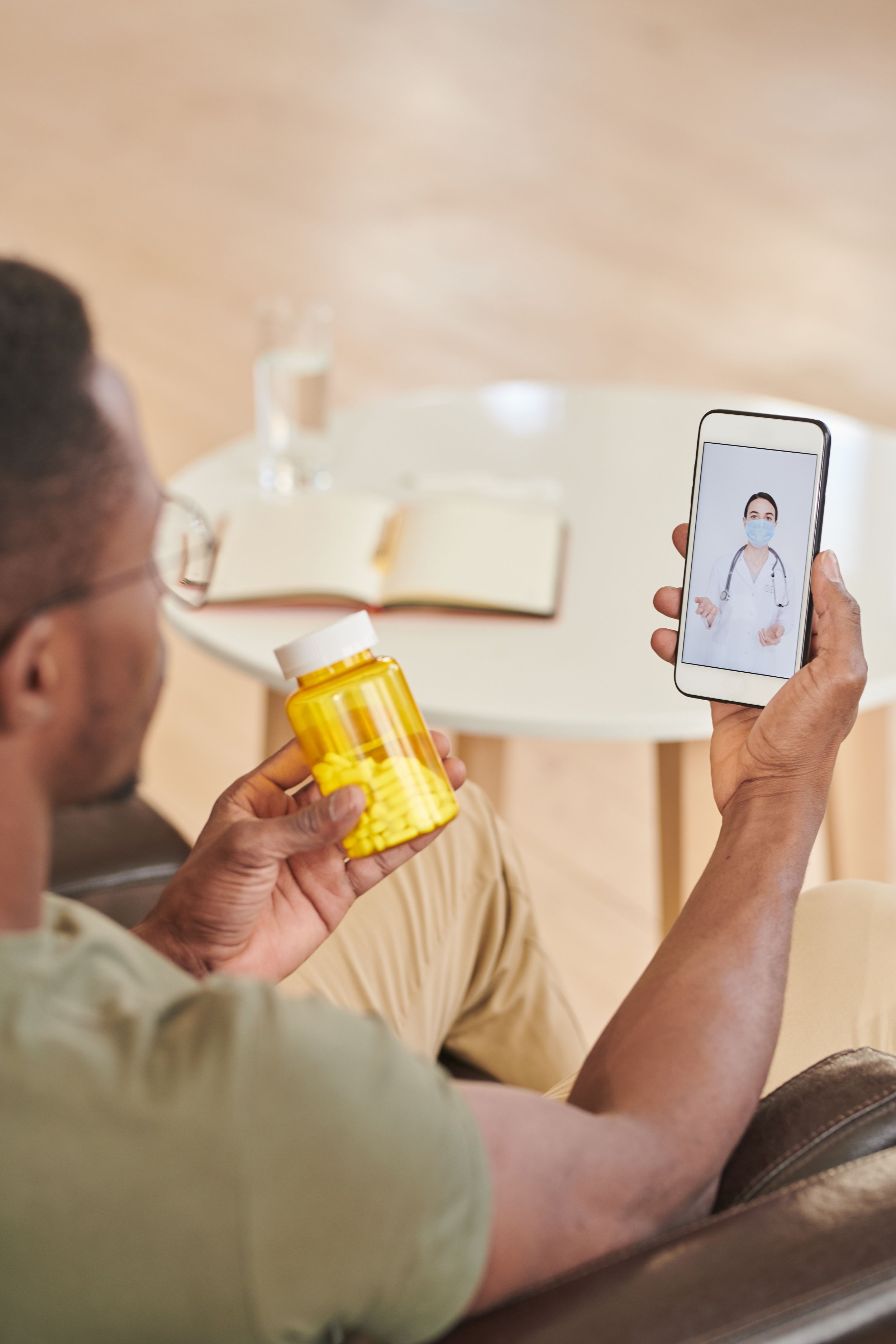 A man holding a bottle of yellow pills looking at a phone displaying a doctor in a mask.