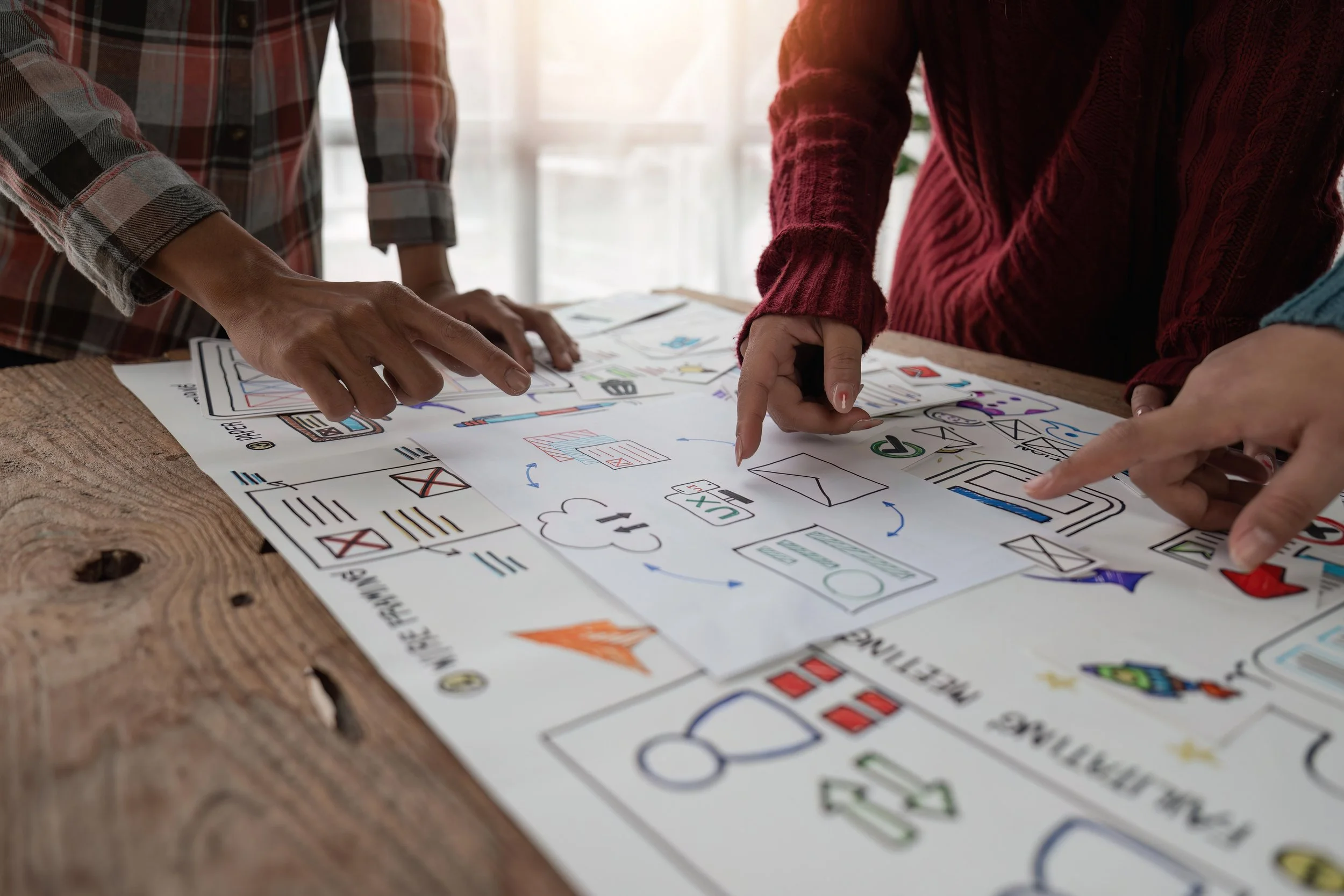 Development team collaborating on a project with sketches and drawings spread out on a wooden table, pointing and discussing.