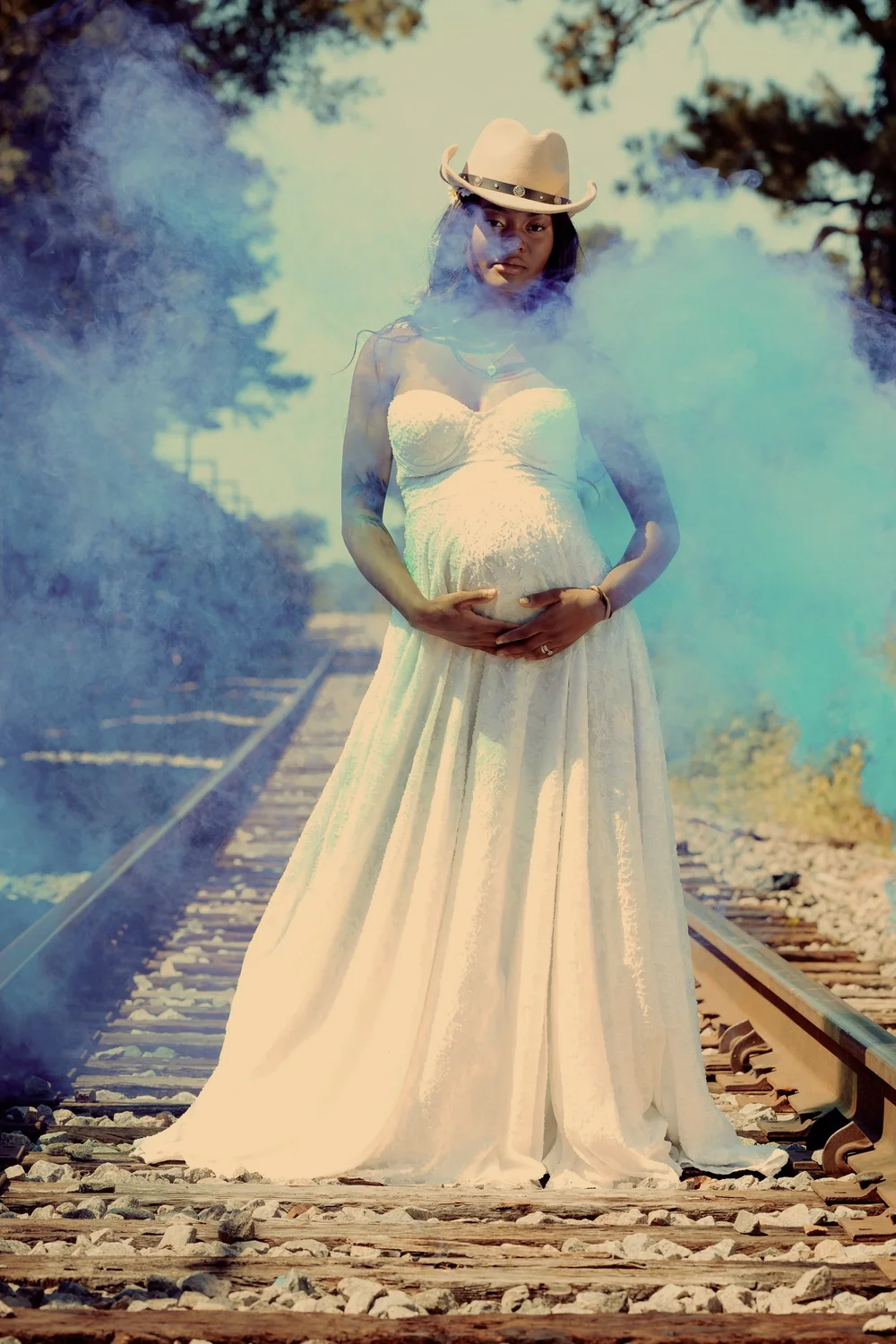 A pregnant woman in a white lace dress and a cowboy hat standing on railway tracks surrounded by blue smoke clouds.