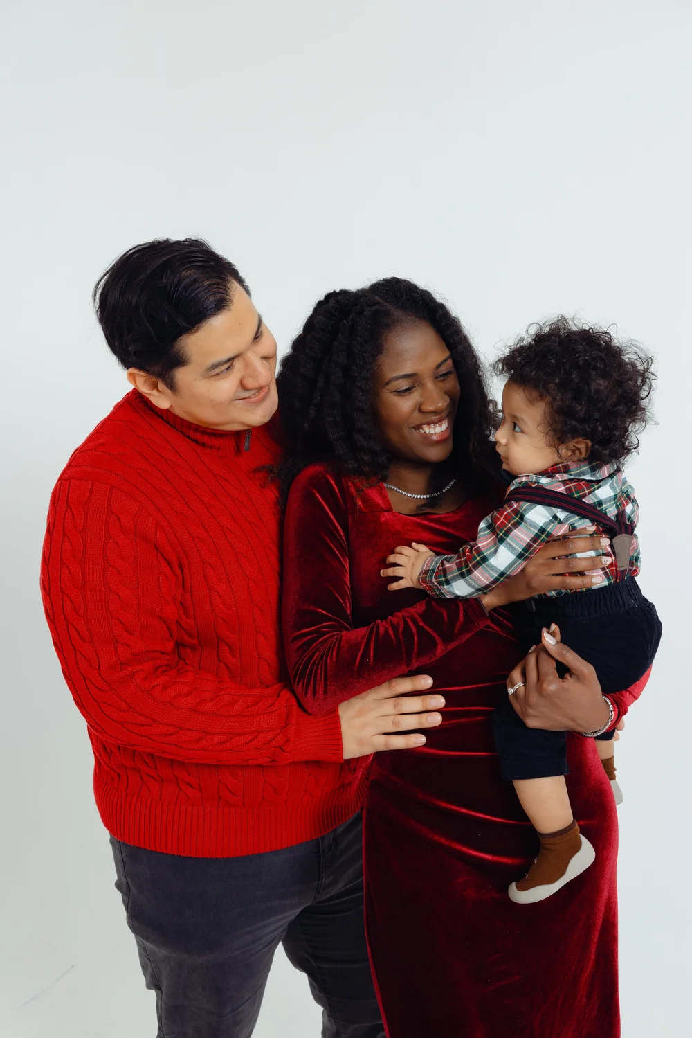 A smiling family with a man in a red sweater, a woman in a red dress, and a toddler in plaid.