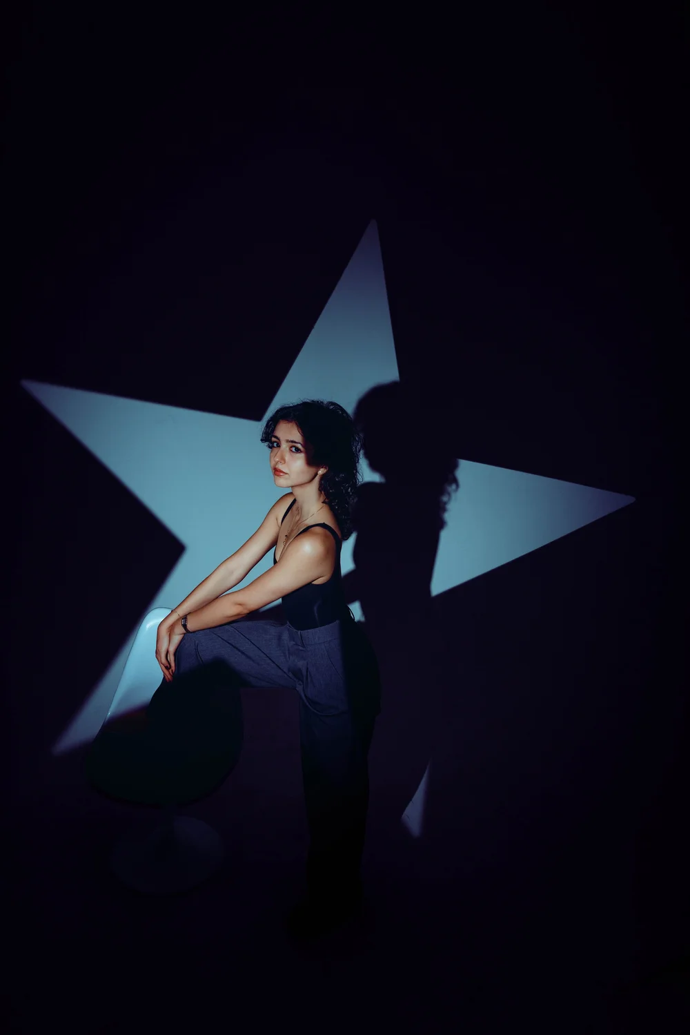 A woman with curly black hair, wearing a black sleeveless top and gray pants, sitting on a white chair in front of a dark background with a large, illuminated star projection, casting a shadow of herself.