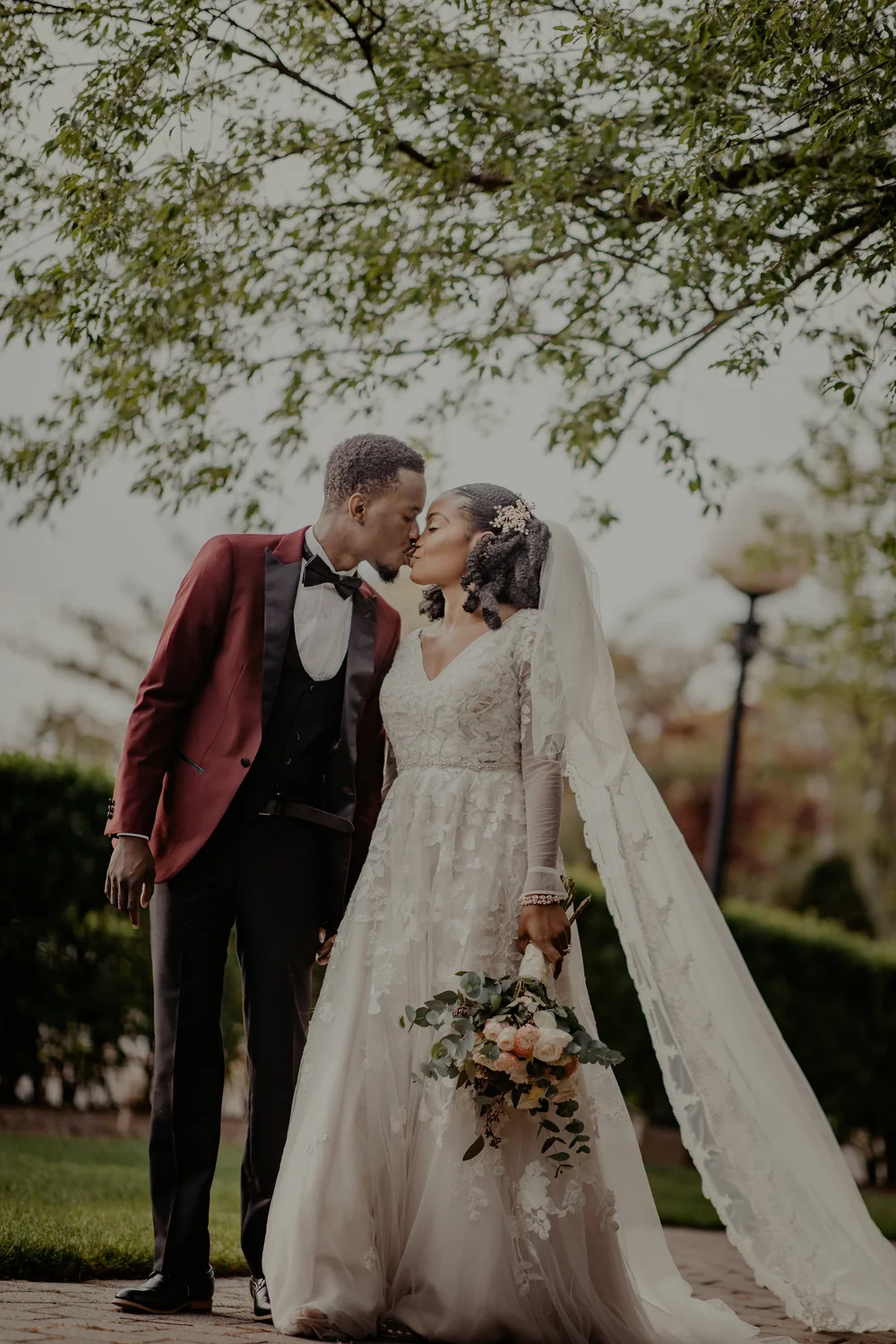 Bride and groom kissing outdoors, wearing wedding attire. Bride holds bouquet. Trees and greenery in background.