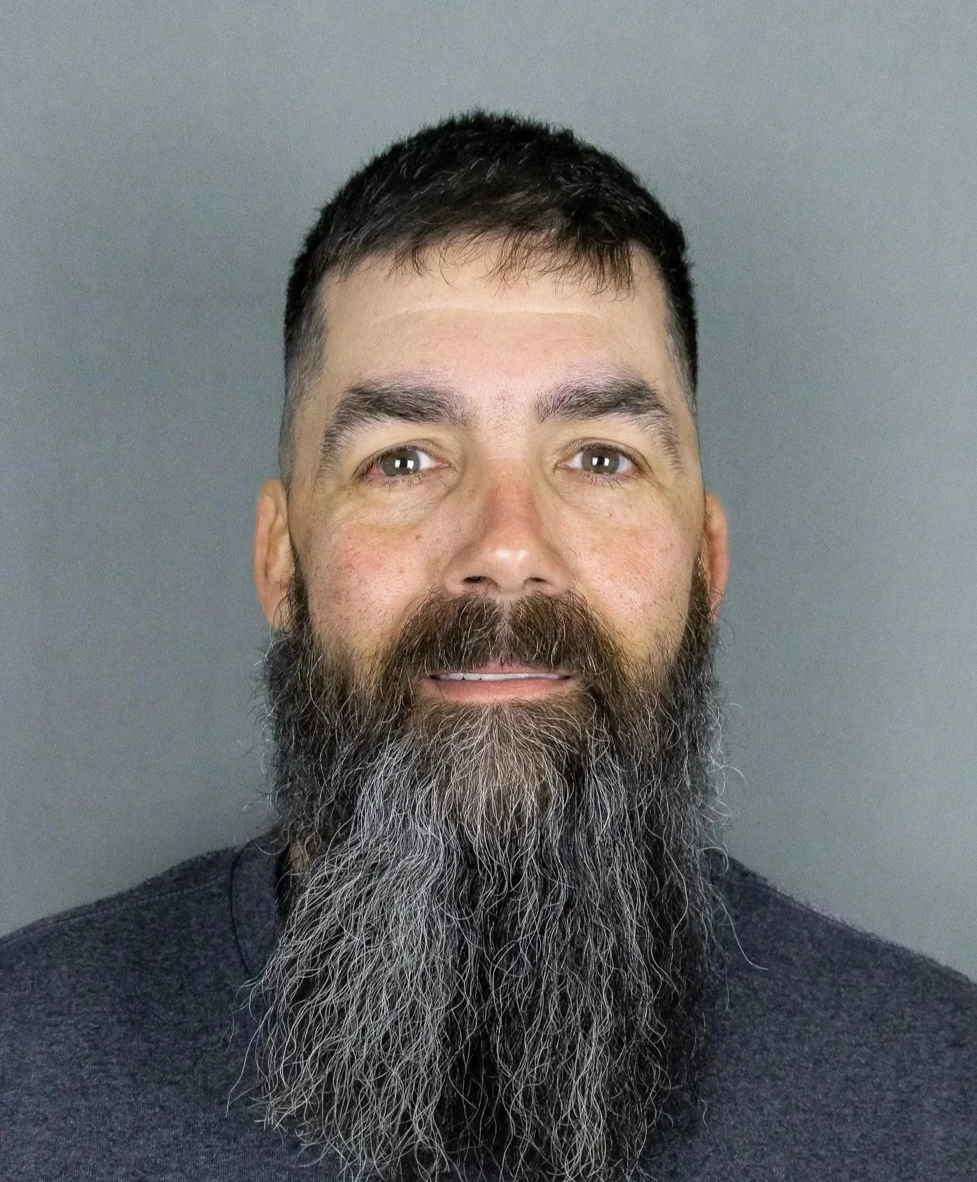 A man with short dark hair and a full beard, wearing a dark shirt, looking directly at the camera against a plain gray background.