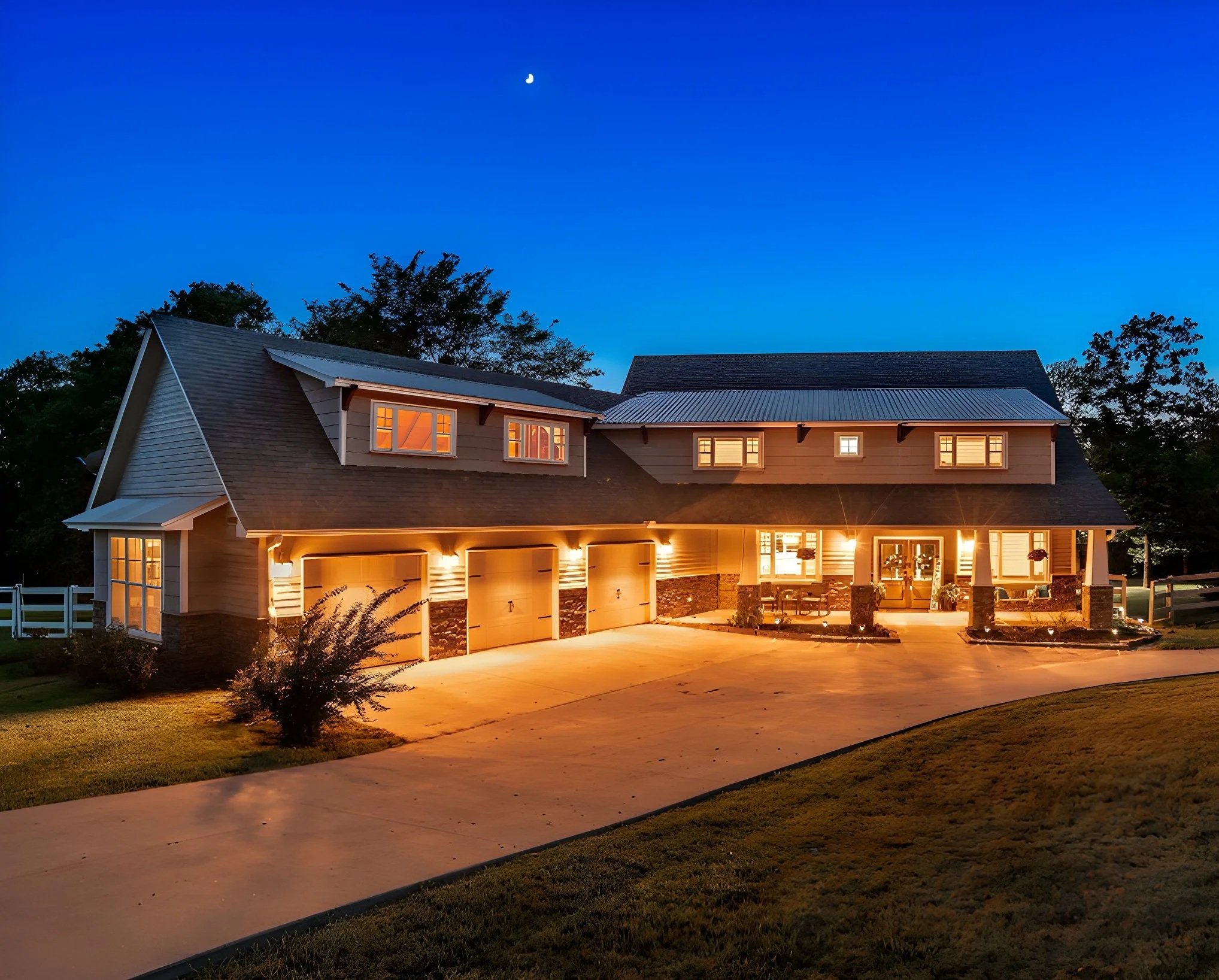 A two-story house with illuminated windows and outdoor lights, white garage doors, and a concrete driveway at dusk with the moon in the sky and trees in the background.