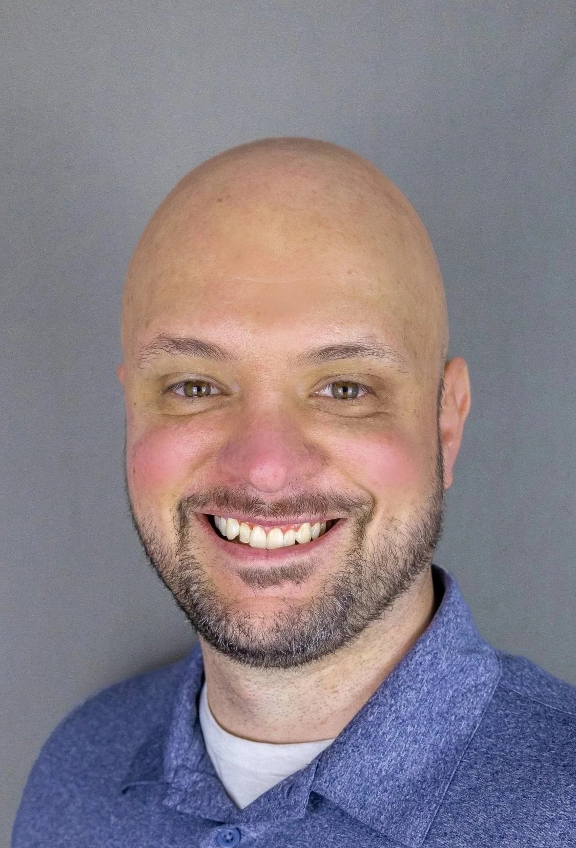 Close-up of a smiling man with a shaved head, beard, wearing a blue collared shirt.