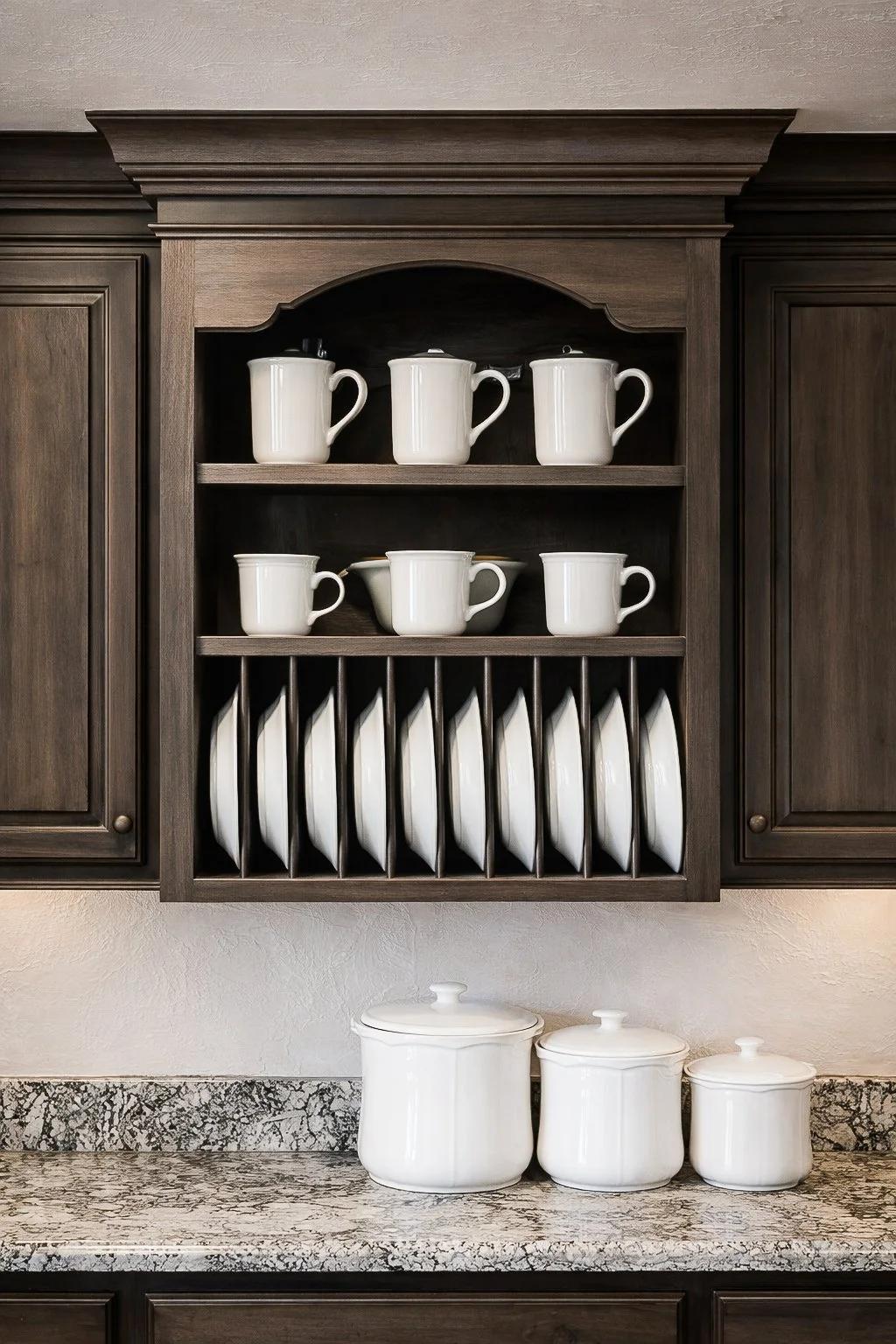 Kitchen cabinet with dark wood finish, holding white mugs, cups, and plates, with three white ceramic storage jars and a granite countertop.