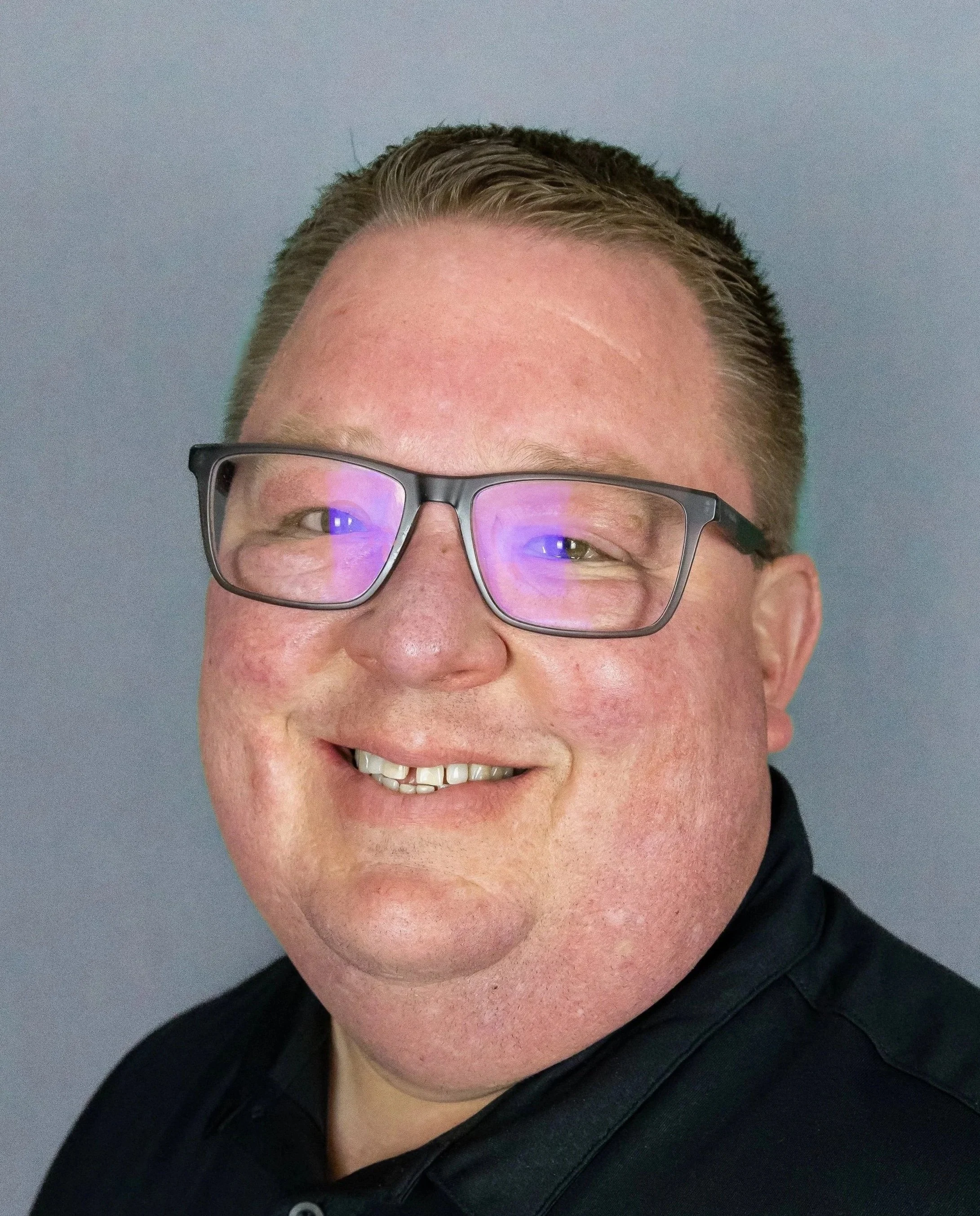 A smiling man with short hair, glasses, and a black shirt, against a plain gray background.