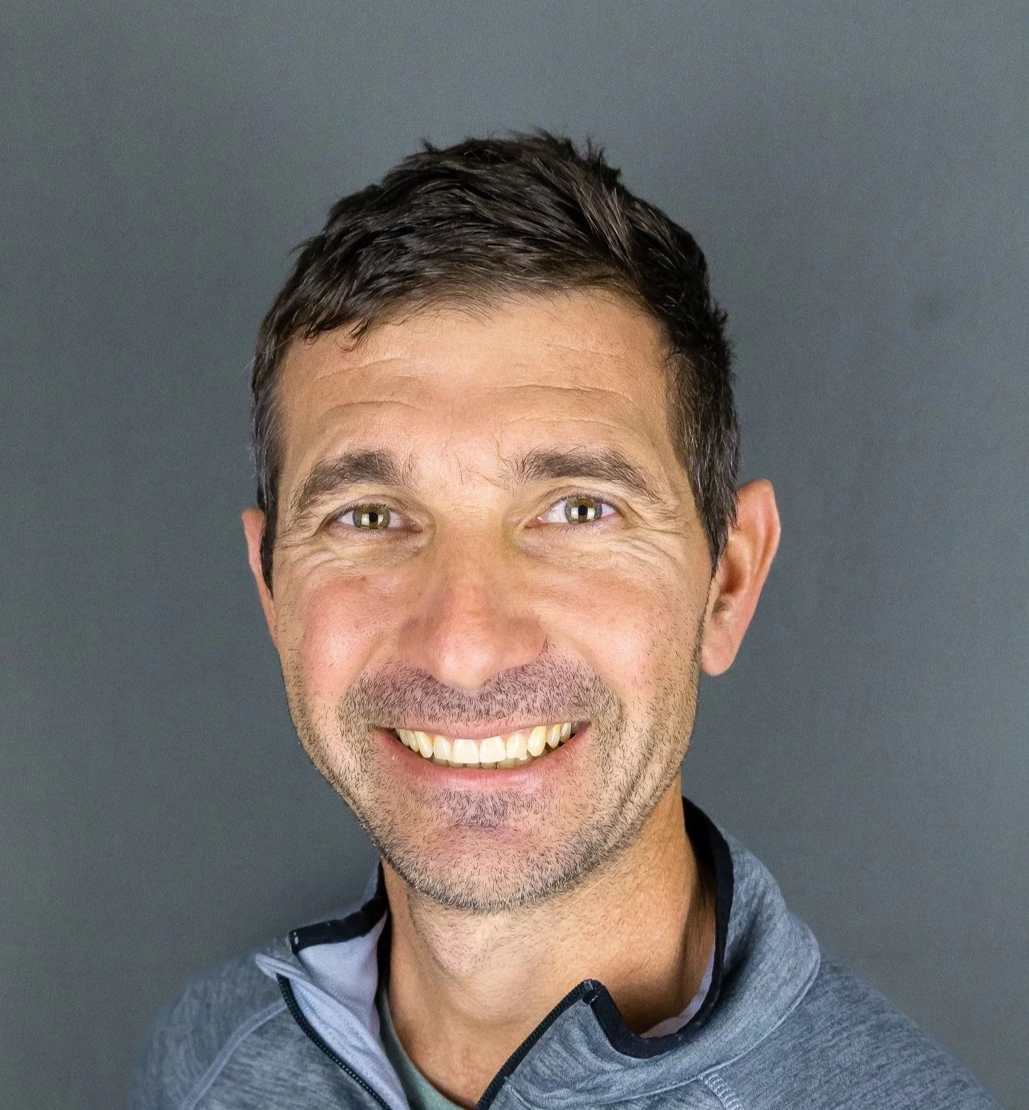 A smiling man with short dark hair and light-colored eyes, wearing a gray athletic jacket, against a gray background.