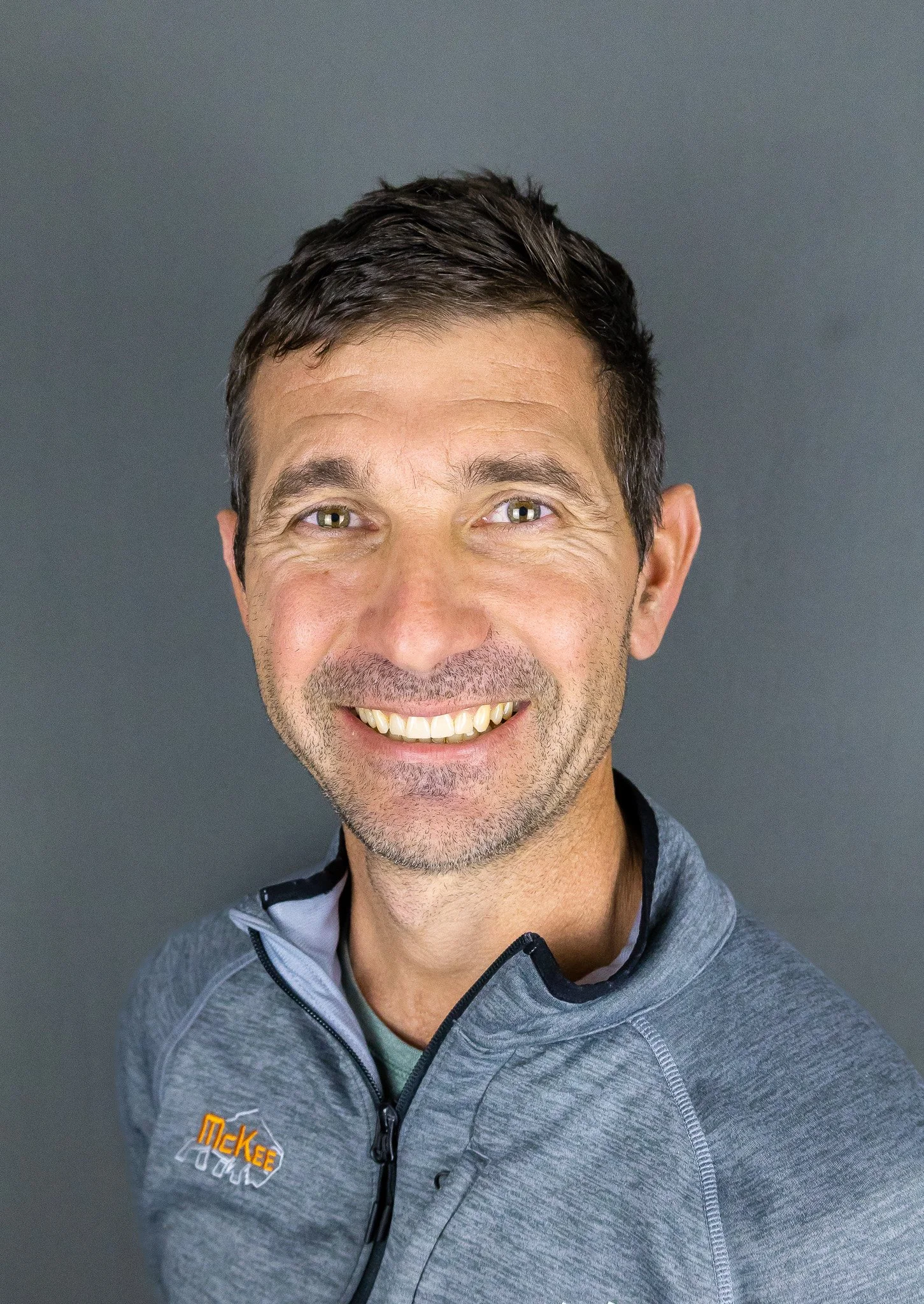 A smiling man with short dark hair and light-colored eyes, wearing a gray zip-up jacket with a small logo on the chest, standing against a gray background.