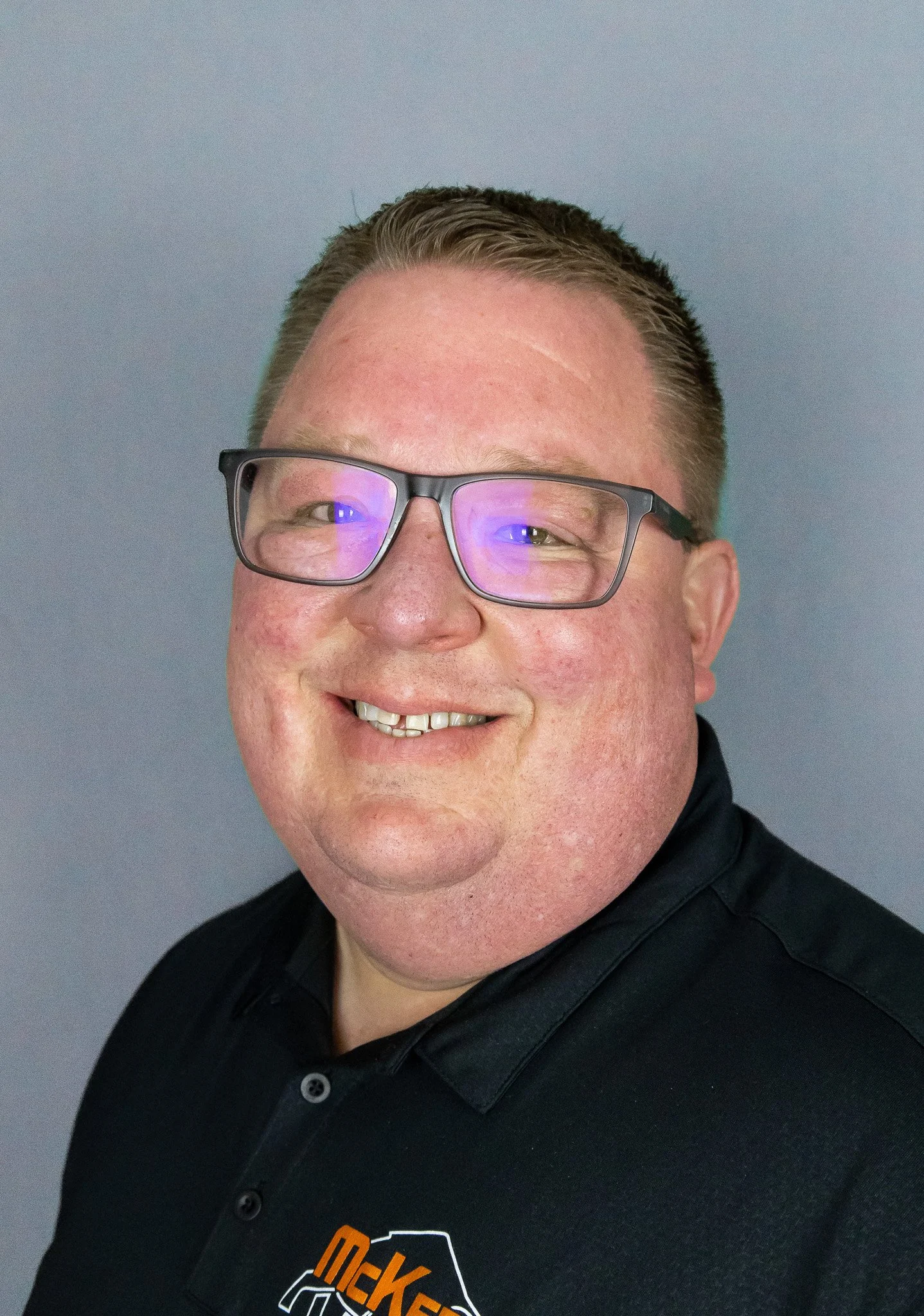 Portrait of a man with short hair, glasses, smiling, wearing a black collared shirt with an orange and white logo.
