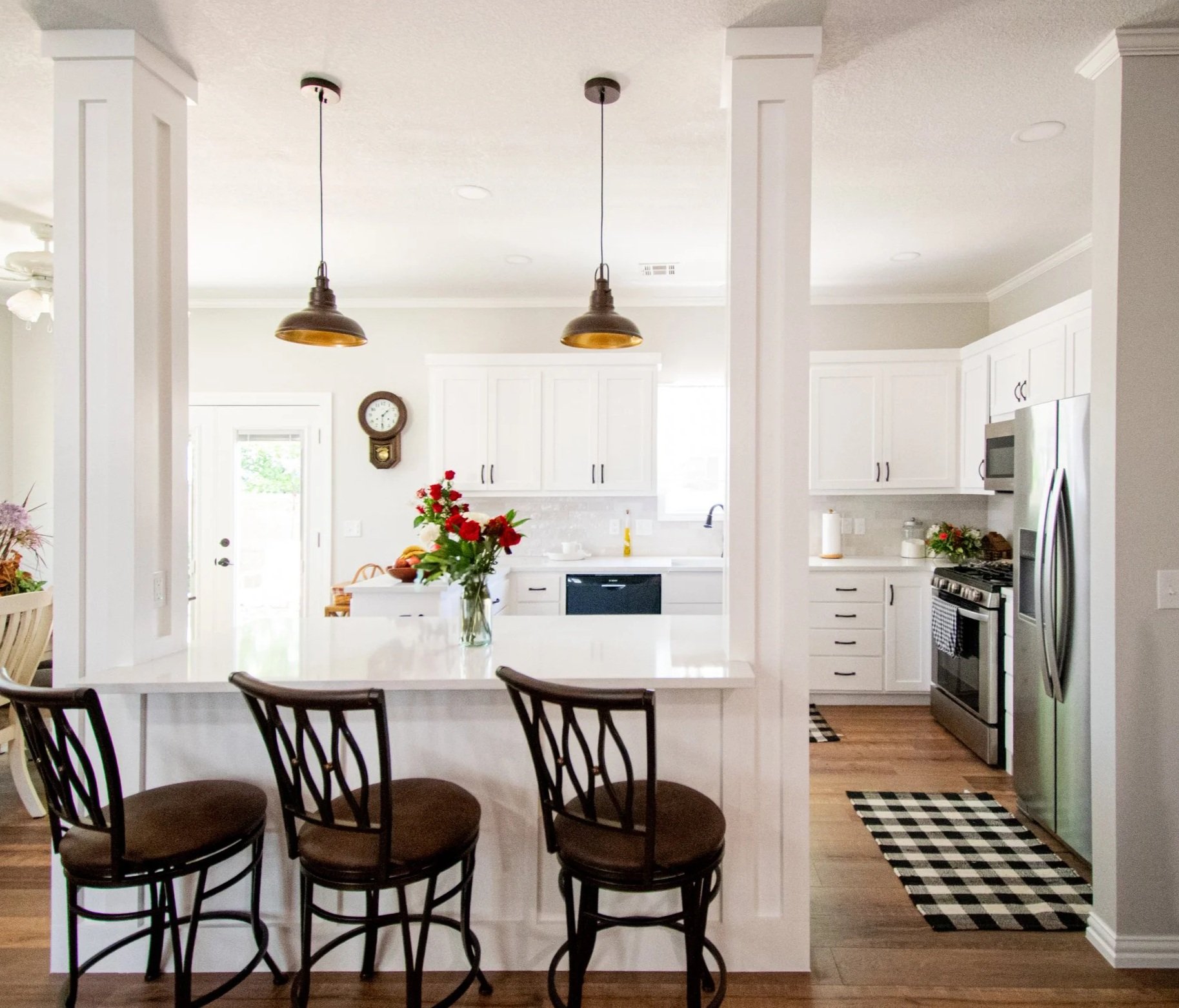 A bright, modern kitchen with white cabinets, a white countertop, and stainless steel appliances. A vase of red flowers sits on the island, which has three black chairs with brown cushions. Two pendant lights hang from the ceiling above the island. The kitchen has a checkered black and white rug on the wooden floor and a wall clock on the back wall.