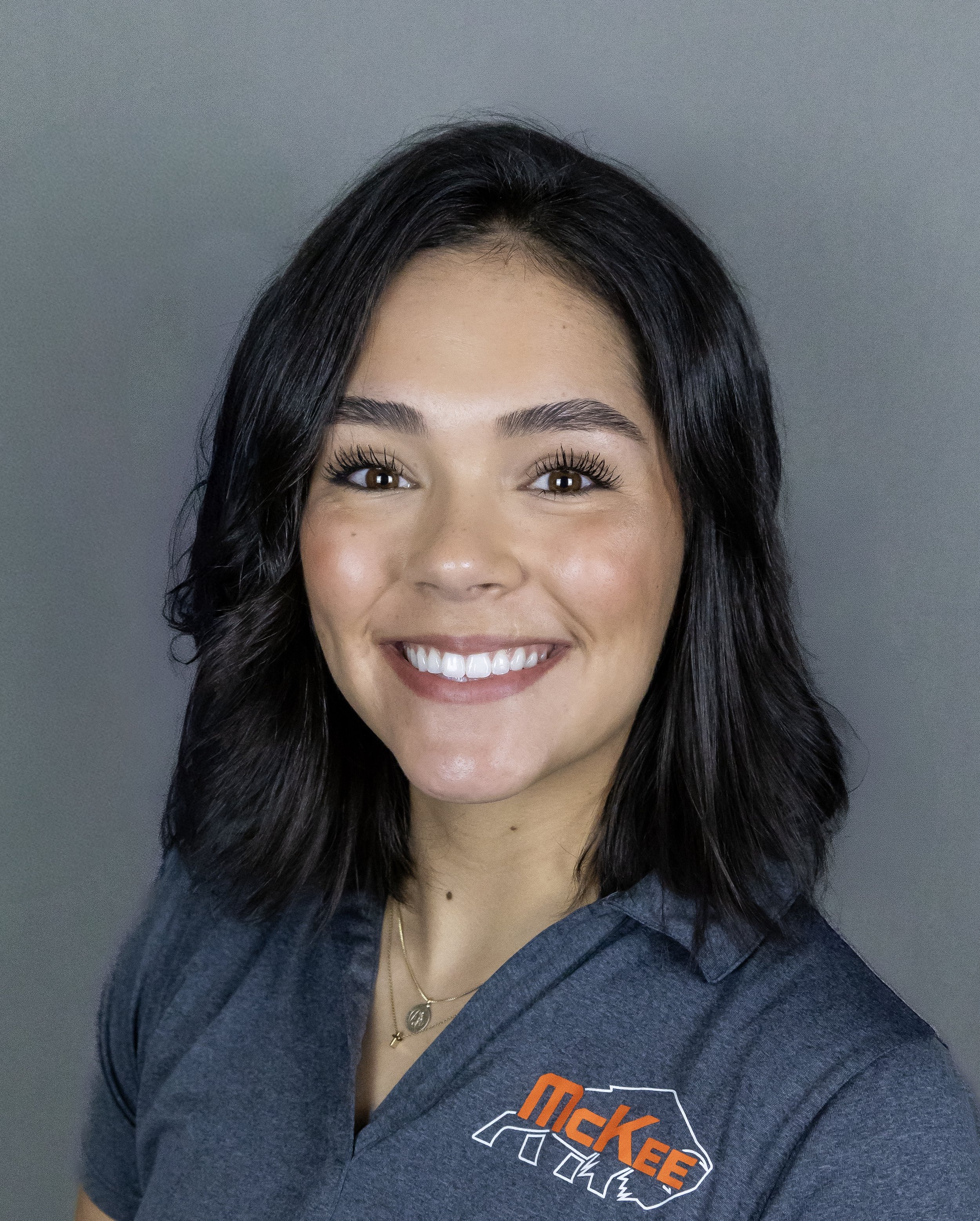A woman with shoulder-length black hair, brown eyes, and a bright smile, wearing a gray polo shirt with the McKee logo, standing against a gray background.