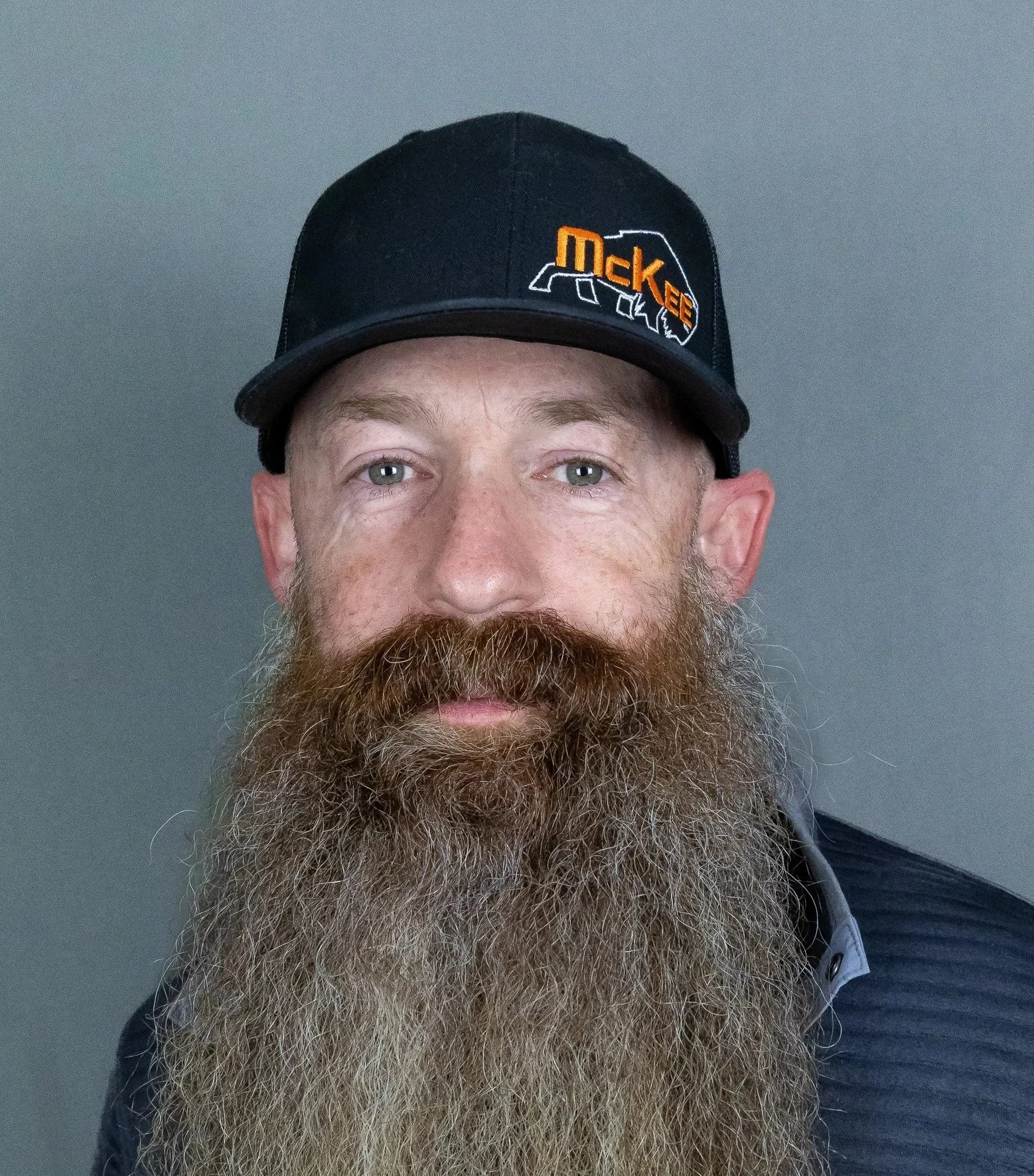 Portrait of a man with a long beard, wearing a black cap with a logo, against a gray background.