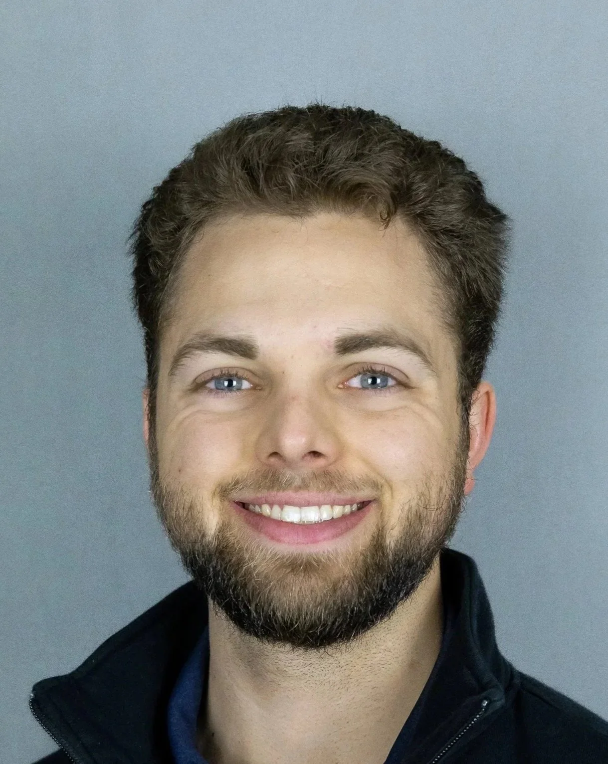 Headshot of a young man with light skin, blue eyes, brown curly hair, and a beard, smiling against a gray background.