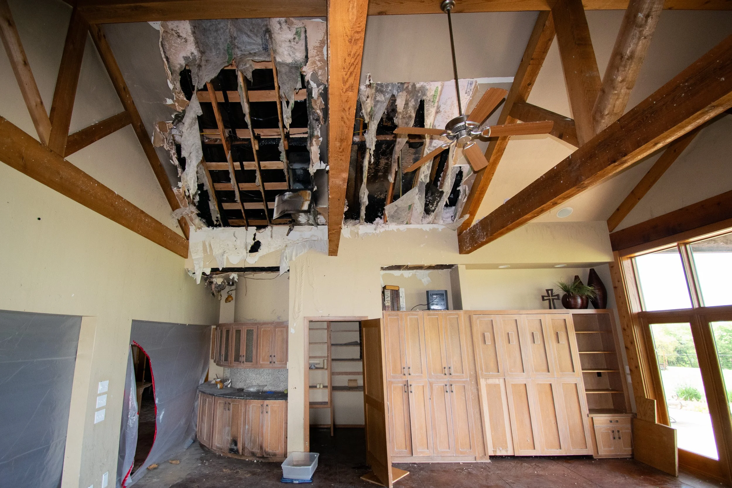 Living room with damaged ceiling exposing ceiling beams, ceiling fan, wooden cabinets, and large window.