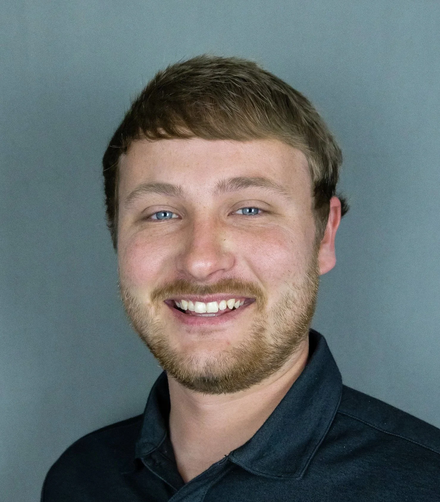 Portrait of a young man with light brown hair, blue eyes, and a beard, smiling, wearing a dark shirt against a gray background.
