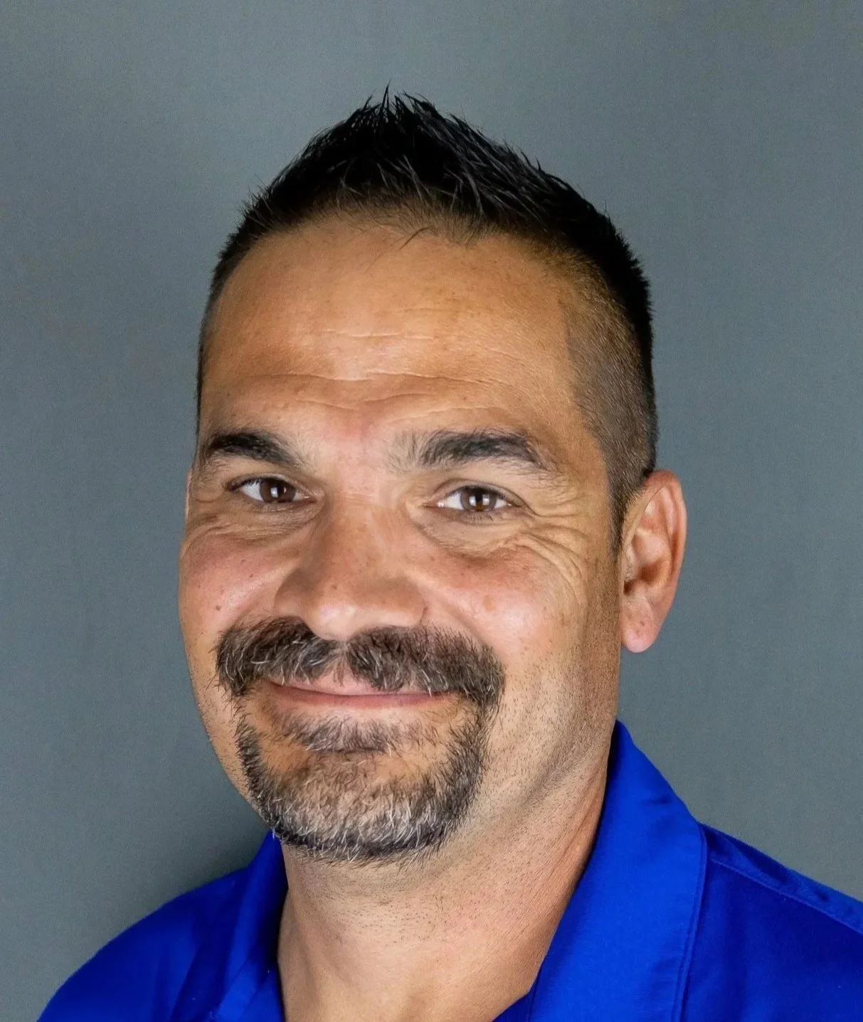 A smiling man with short dark hair and a goatee, wearing a blue shirt, standing against a gray background.