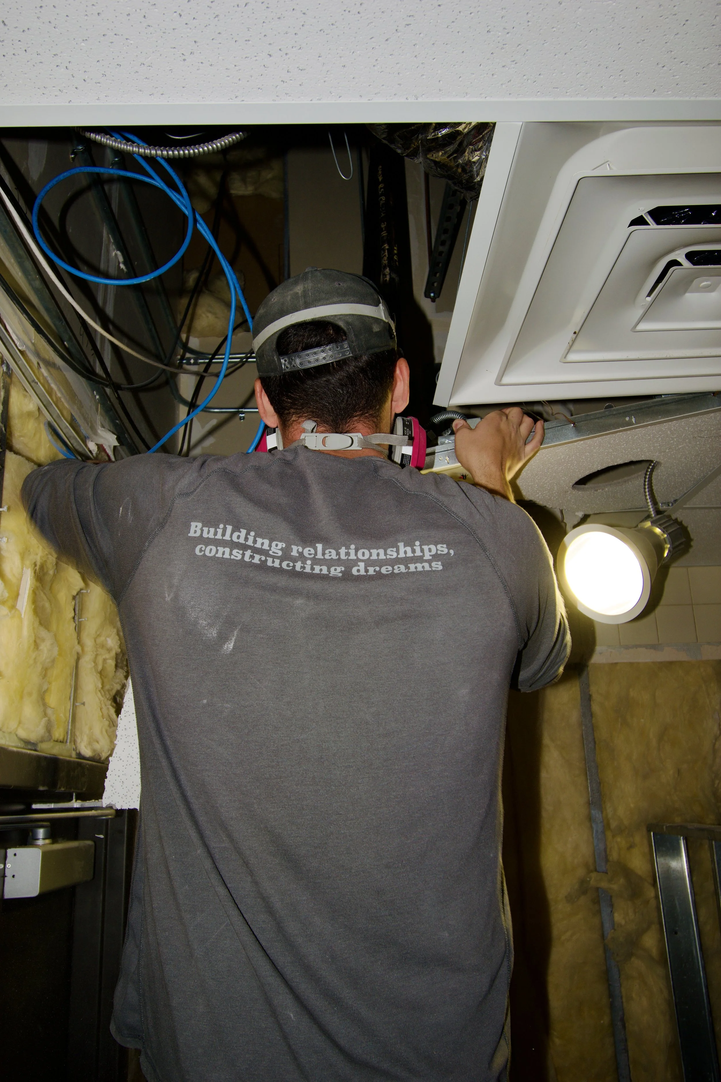 A man working on an HVAC system installation or repair, wearing a cap and a t-shirt that says 'Building relationships, constructing dreams' in a construction or maintenance setting.