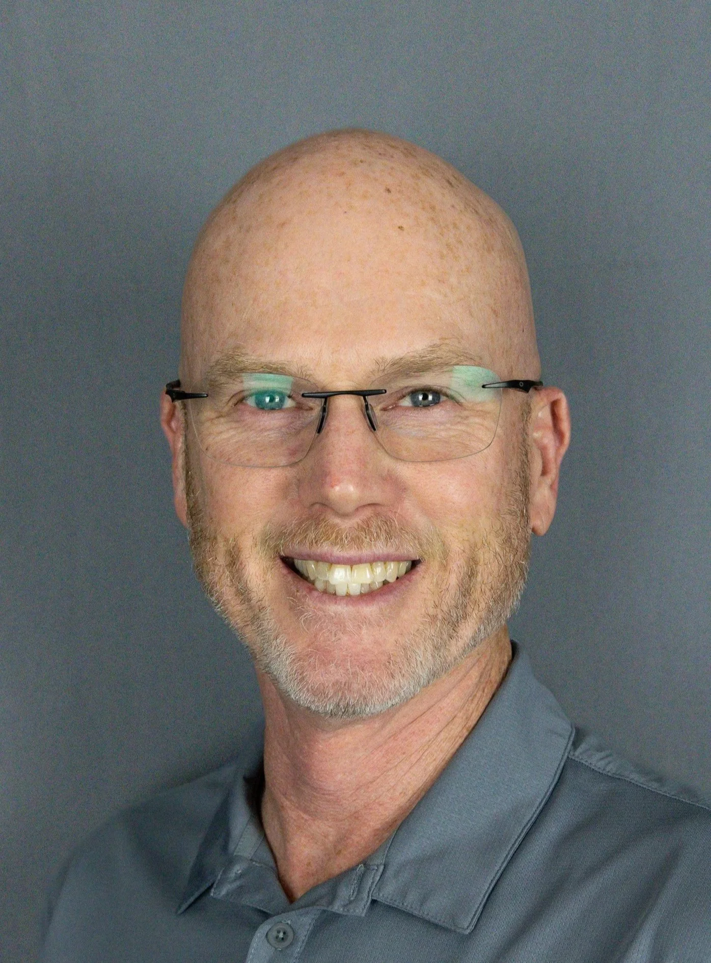 A smiling man with a shaved head, glasses, and a beard, wearing a gray collared shirt, against a gray background.