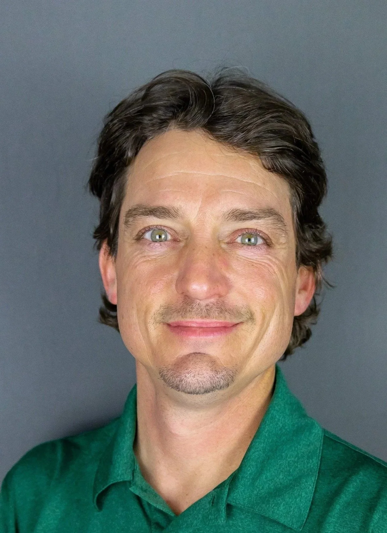 A man with dark, wavy hair and blue eyes, smiling, wearing a green collared shirt, standing against a plain, dark gray background.