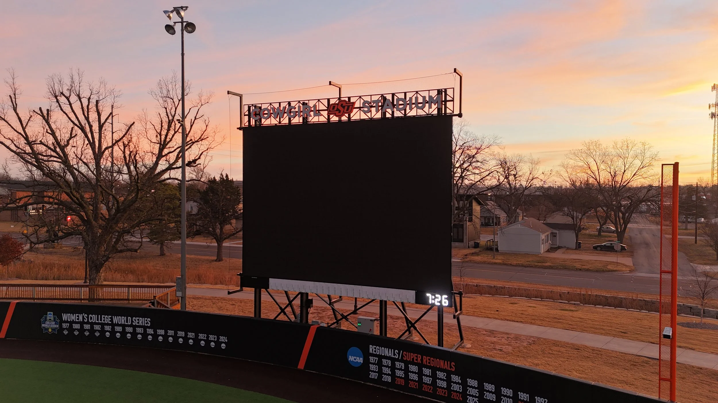 A large, empty outdoor stadium scoreboard at sunset with a banner reading '#COWGIRL OSU STADIUM' on top. There are trees and houses in the background and a small digital clock displaying 7:26. A black banner at the bottom lists NCAA women's college world series and regional/super regionals history.