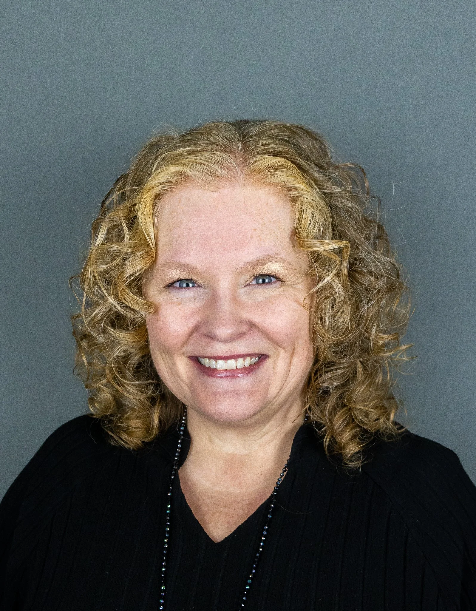 A woman with curly blonde hair and blue eyes, smiling, wearing a black top and a beaded necklace, against a gray background.