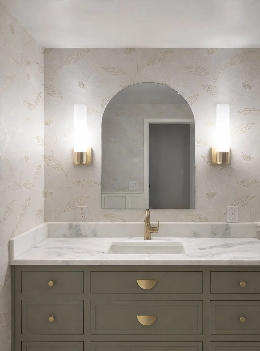 Bathroom vanity with a marble countertop, a gold faucet, and a large mirror framed with floral wallpaper. Two wall-mounted lights are on either side of the mirror.