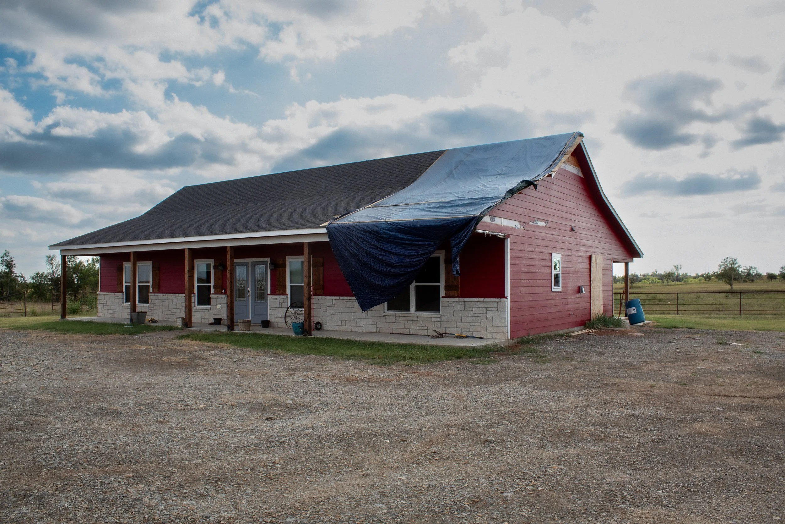 A house under construction with a partial tarp covering the front porch. The house has red siding, white stone accents near the foundation, and a dark gray roof. There are lawn supplies and a blue barrel on the yard, with a rural landscape and partly cloudy sky in the background.