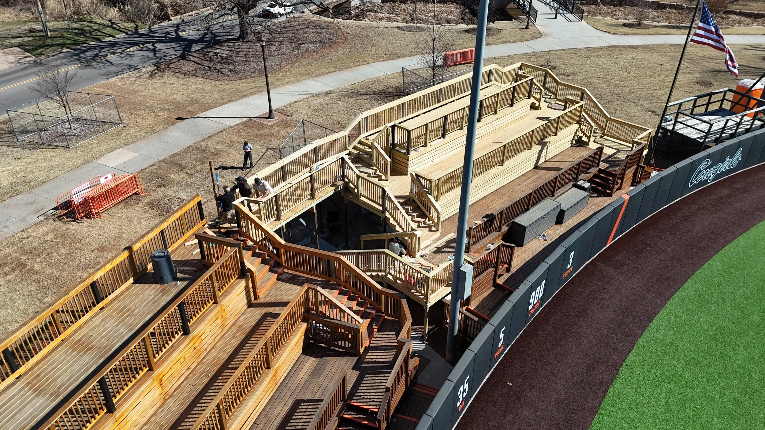 Construction of wooden bleachers at a baseball or sports stadium, with workers installing the seating area, an American flag, and a baseball foul line in the foreground.