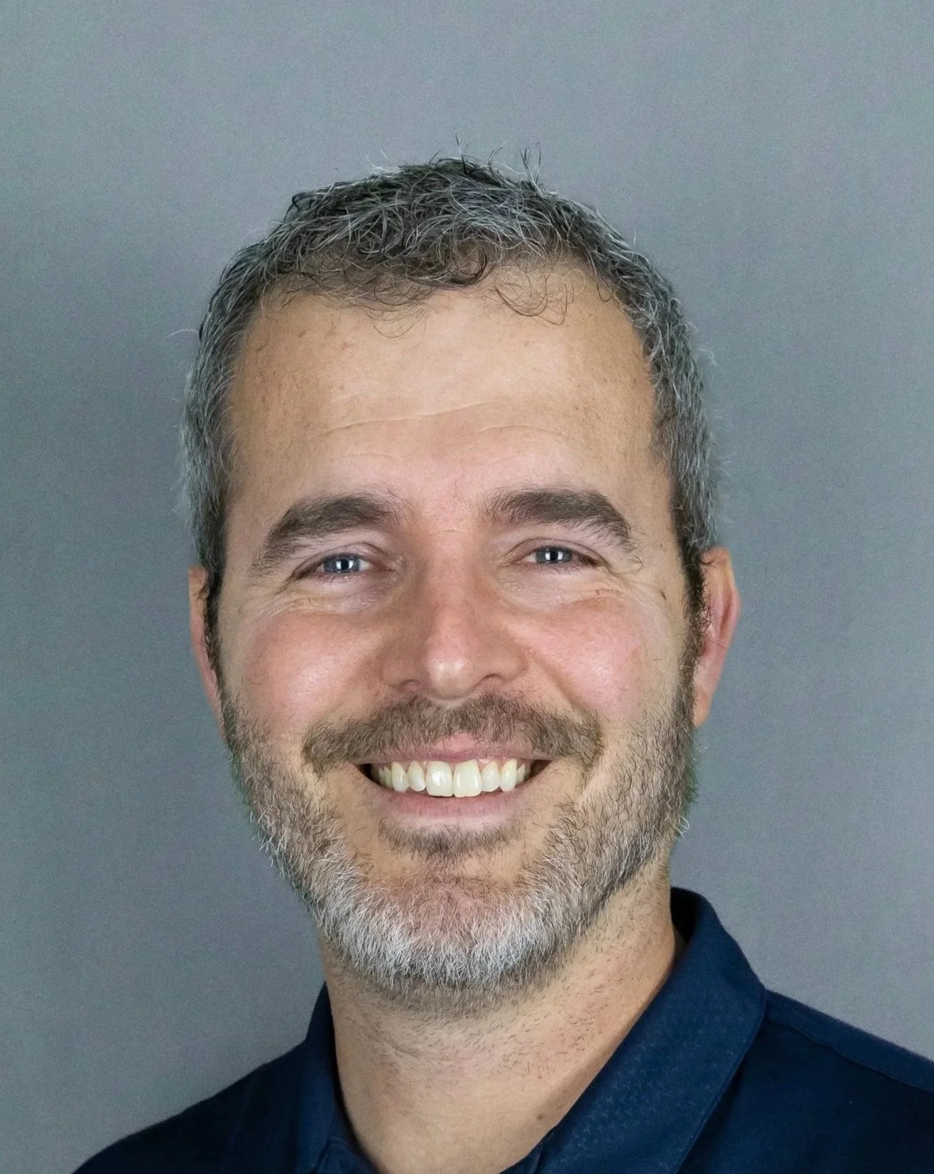 Close-up portrait of a man with short, curly, salt-and-pepper hair and a beard, smiling, wearing a dark blue shirt, against a plain gray background.