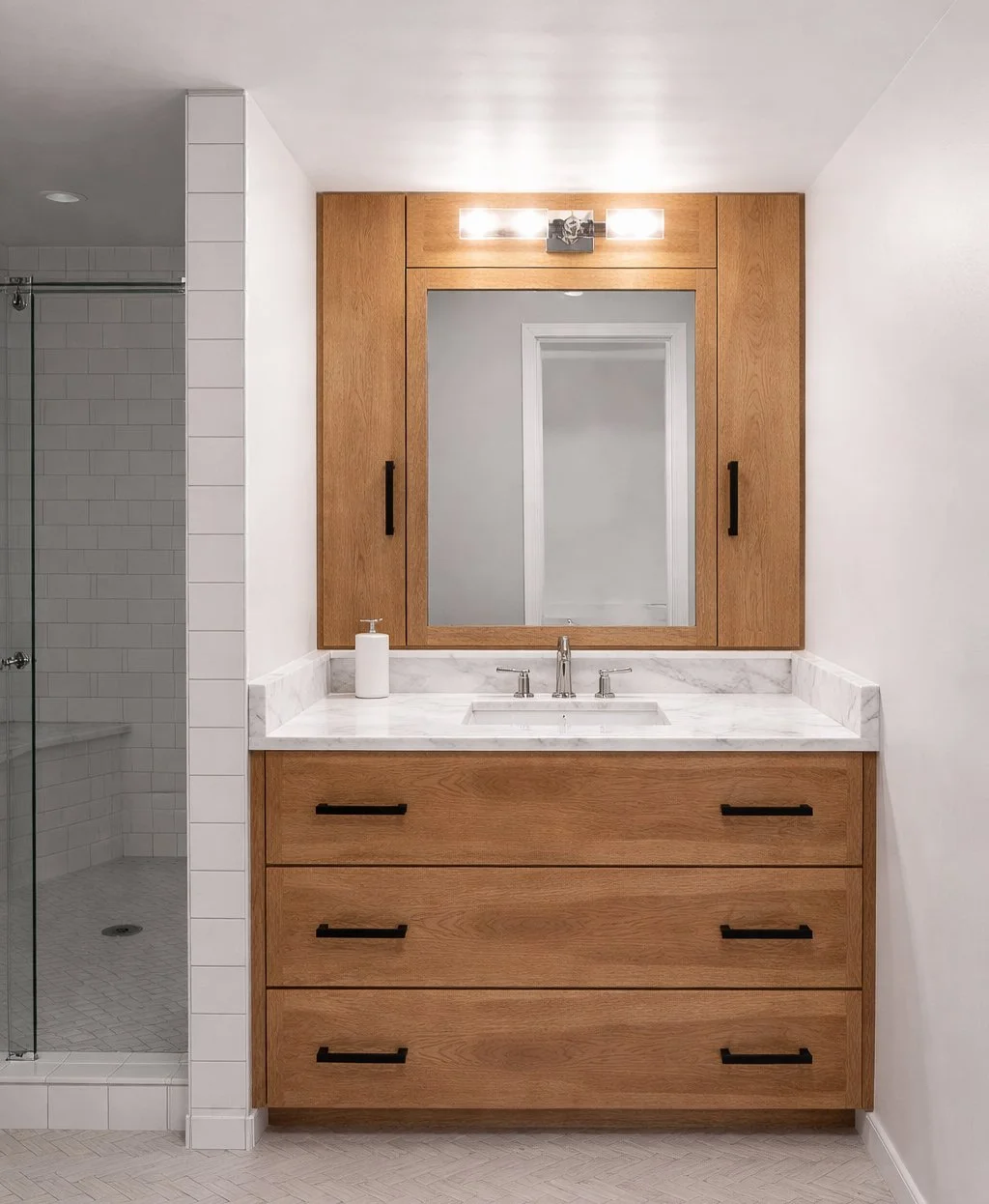 Modern bathroom vanity with a large wooden framed mirror, marble countertop, and four drawers with black handles. A soap dispenser is on the left side of the countertop. To the left, part of a shower with white tiles and a glass door is visible.