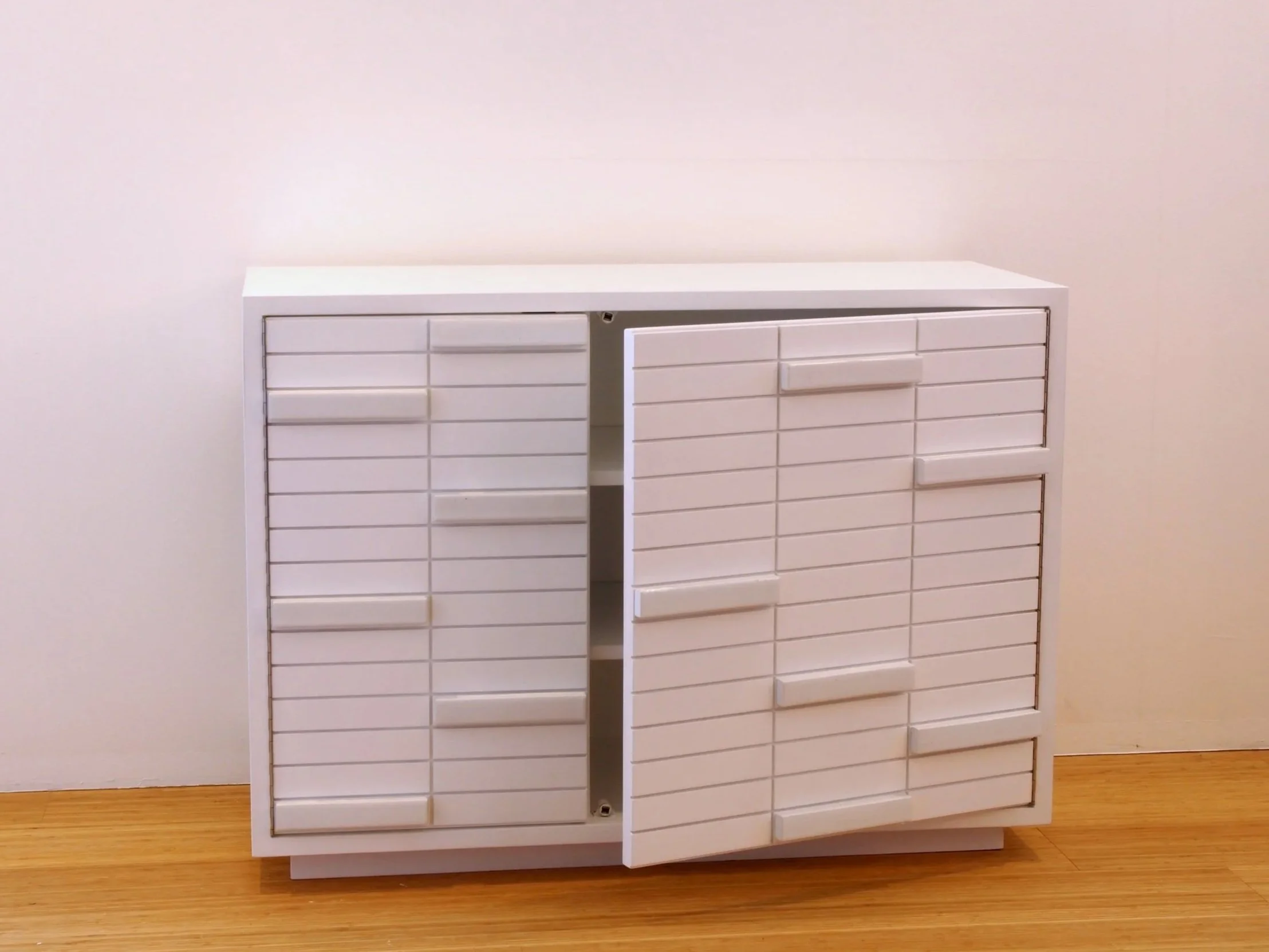 Una cassettiera bianca con molte piccole ante di plastica, alcune delle quali sono aperte, appoggiata su un pavimento di legno, contro una parete bianca.