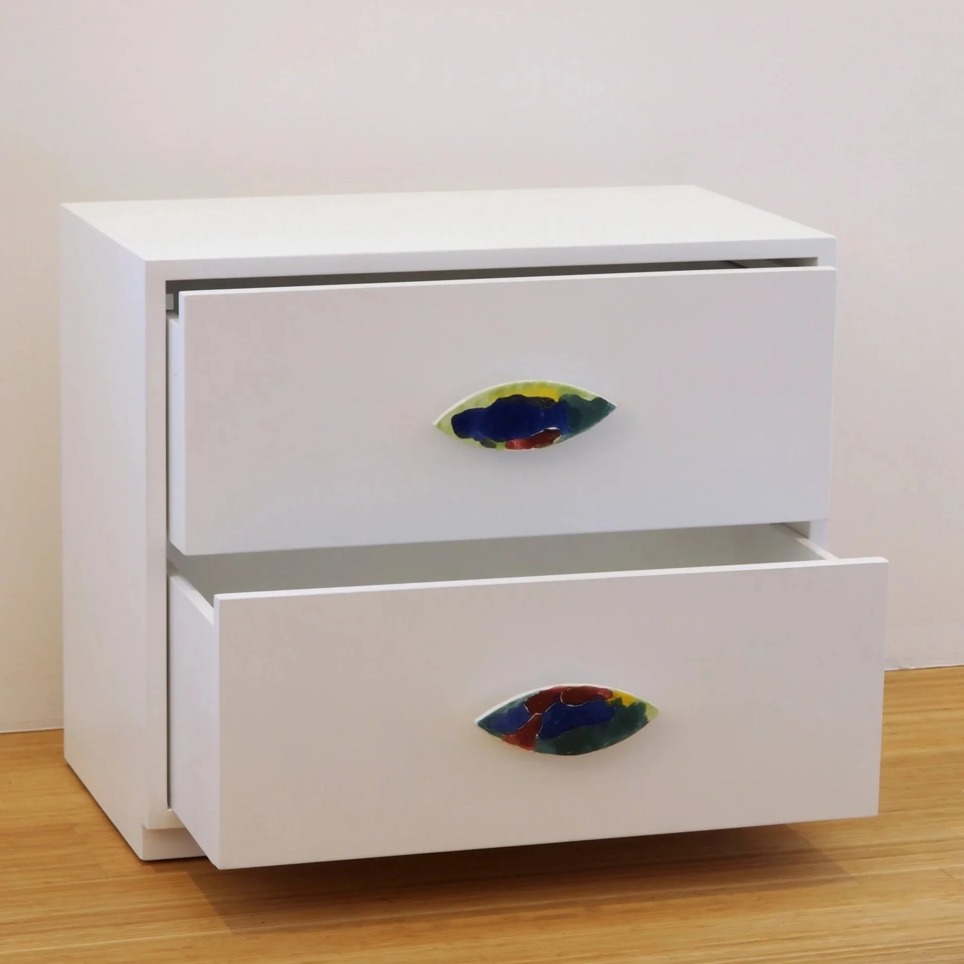 Cassettiera bianca con due cassetti, ciascuno con un pomello in ceramica decorativo colorato a forma di occhio stilizzato.