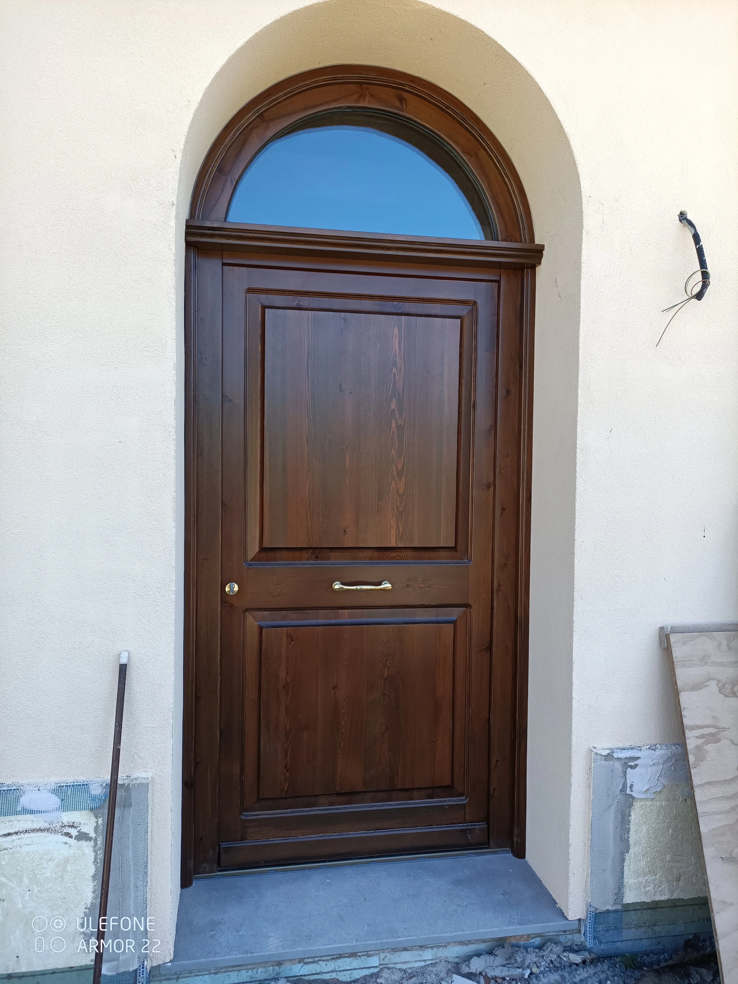 Porta in legno con arco sopra, installata di fronte a una parete chiara, con un marco di pietra o cemento davanti.