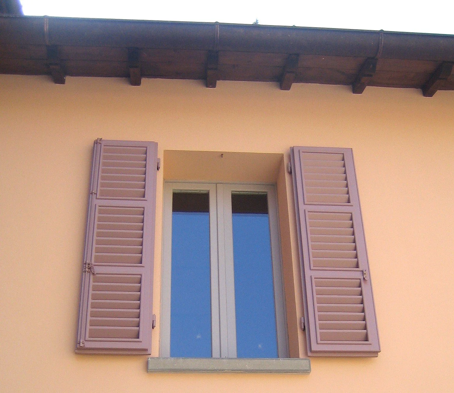 Finestra con persiane di colore beige su parete di colore giallo, con tettuccio di legno sopra.