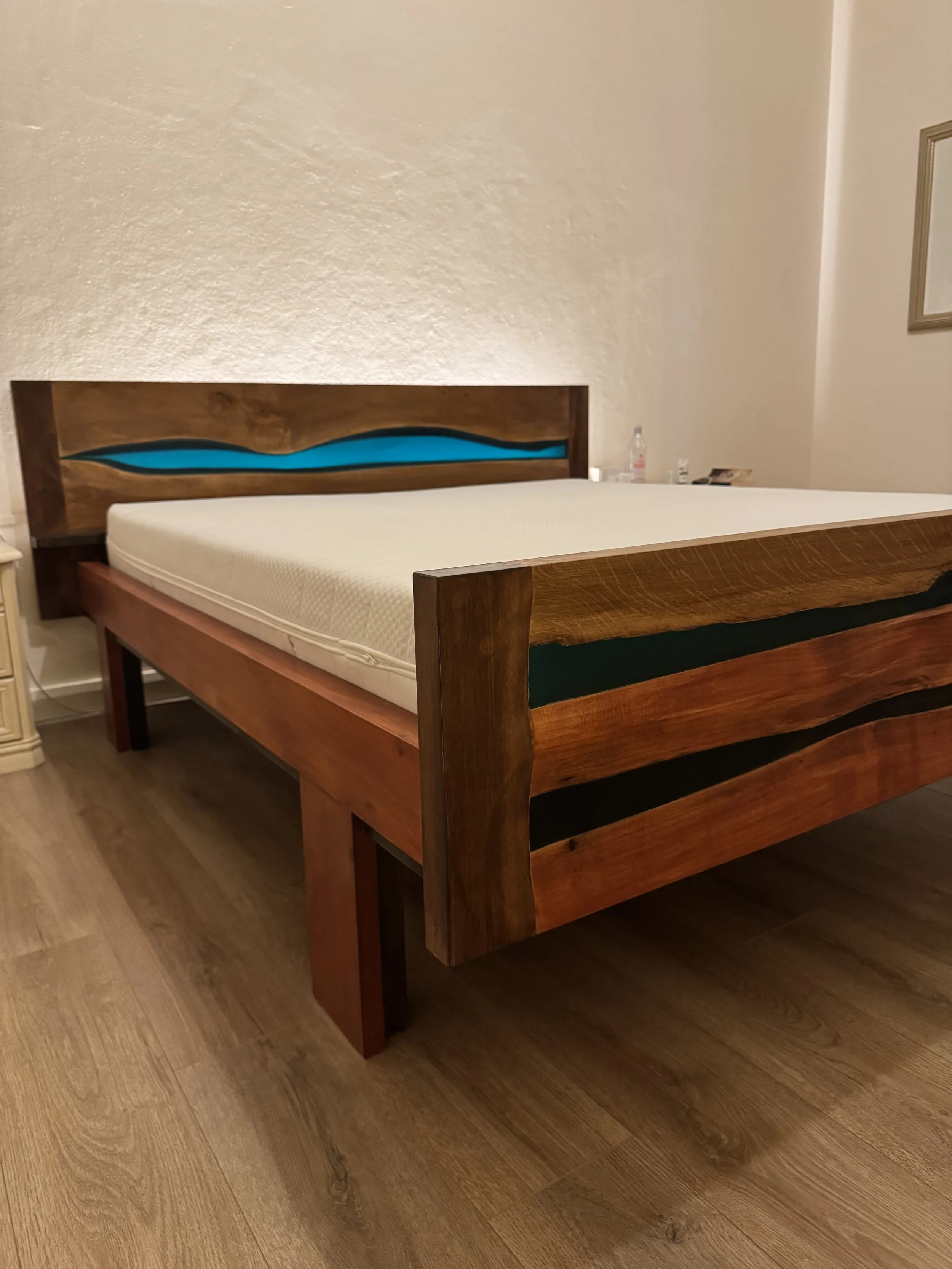 Letto matrimoniale in legno con testata decorata da un intaglio con elemento blu, in una stanza con pareti beige e pavimento in legno.